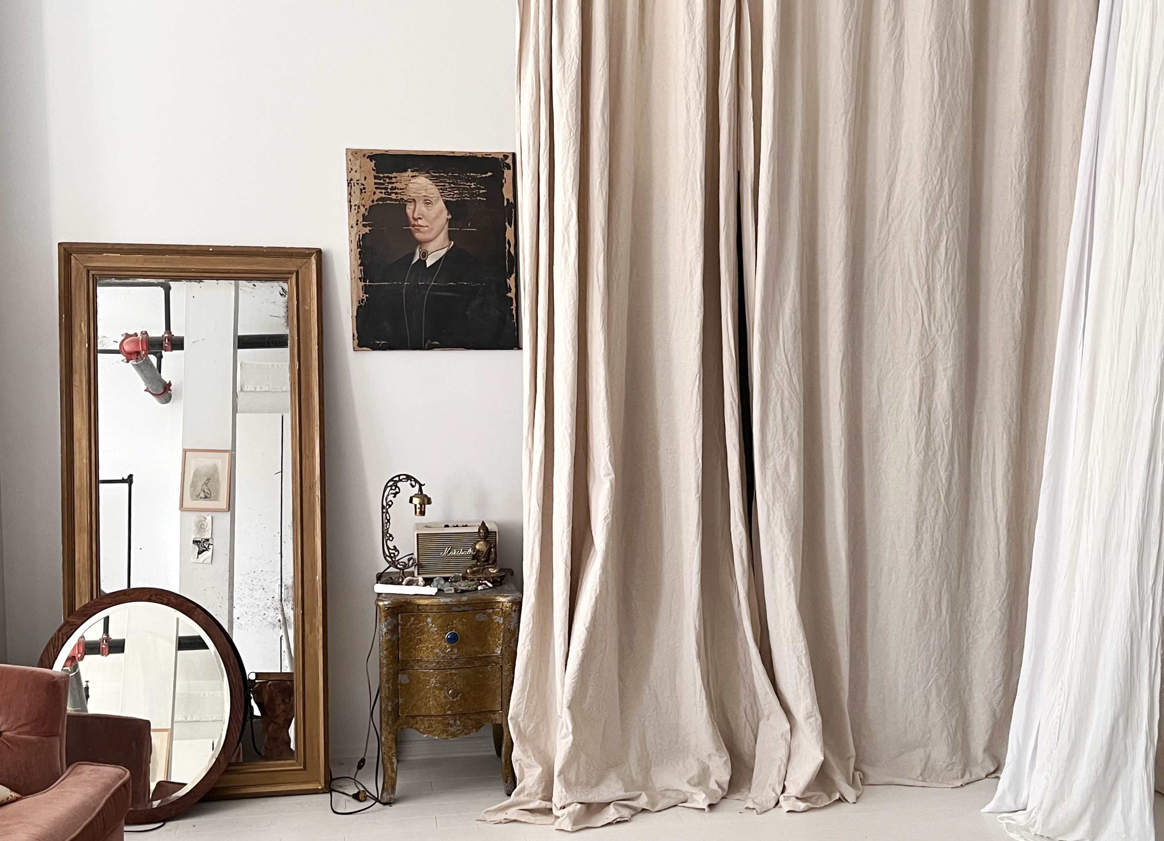 A room with a large mirror, a framed painting, beige drapes, a wooden table with decorative items, and a sofa in the foreground.