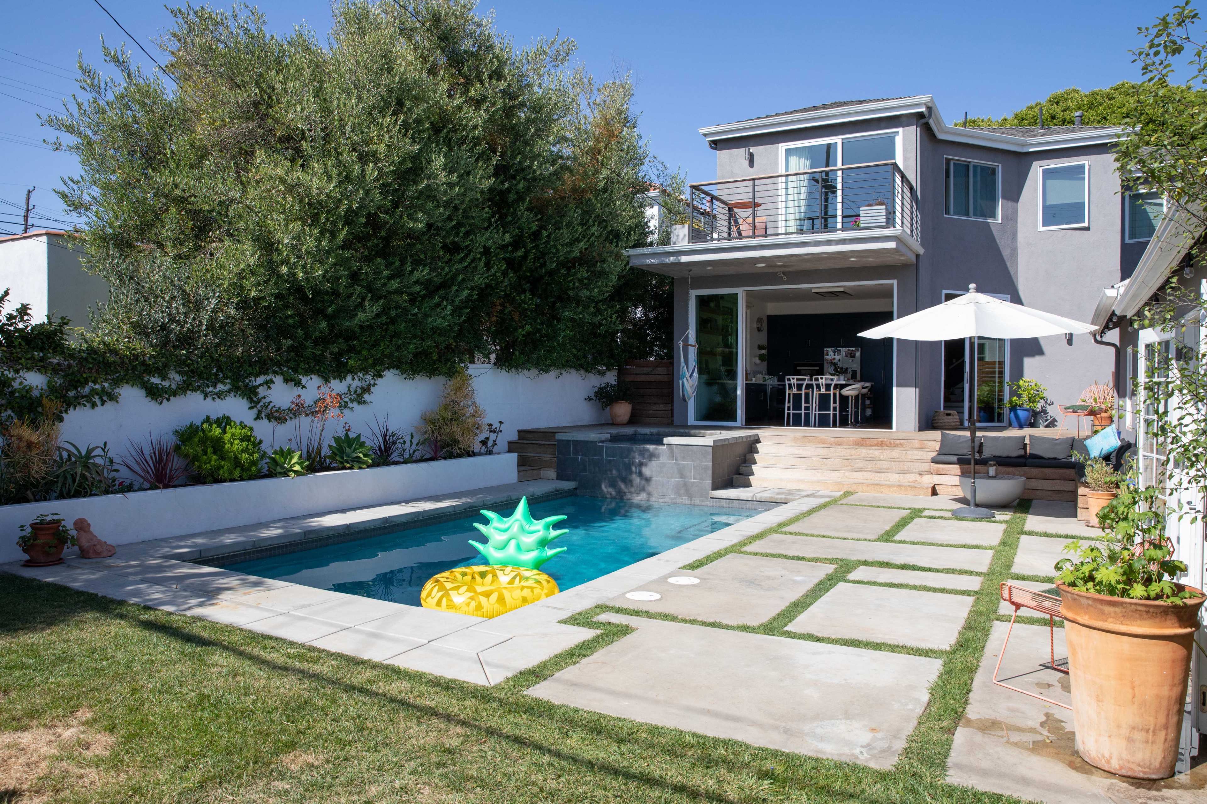 The image shows a modern backyard featuring a pool with a pineapple float, surrounded by concrete walkways, grass, and a patio with outdoor furniture.