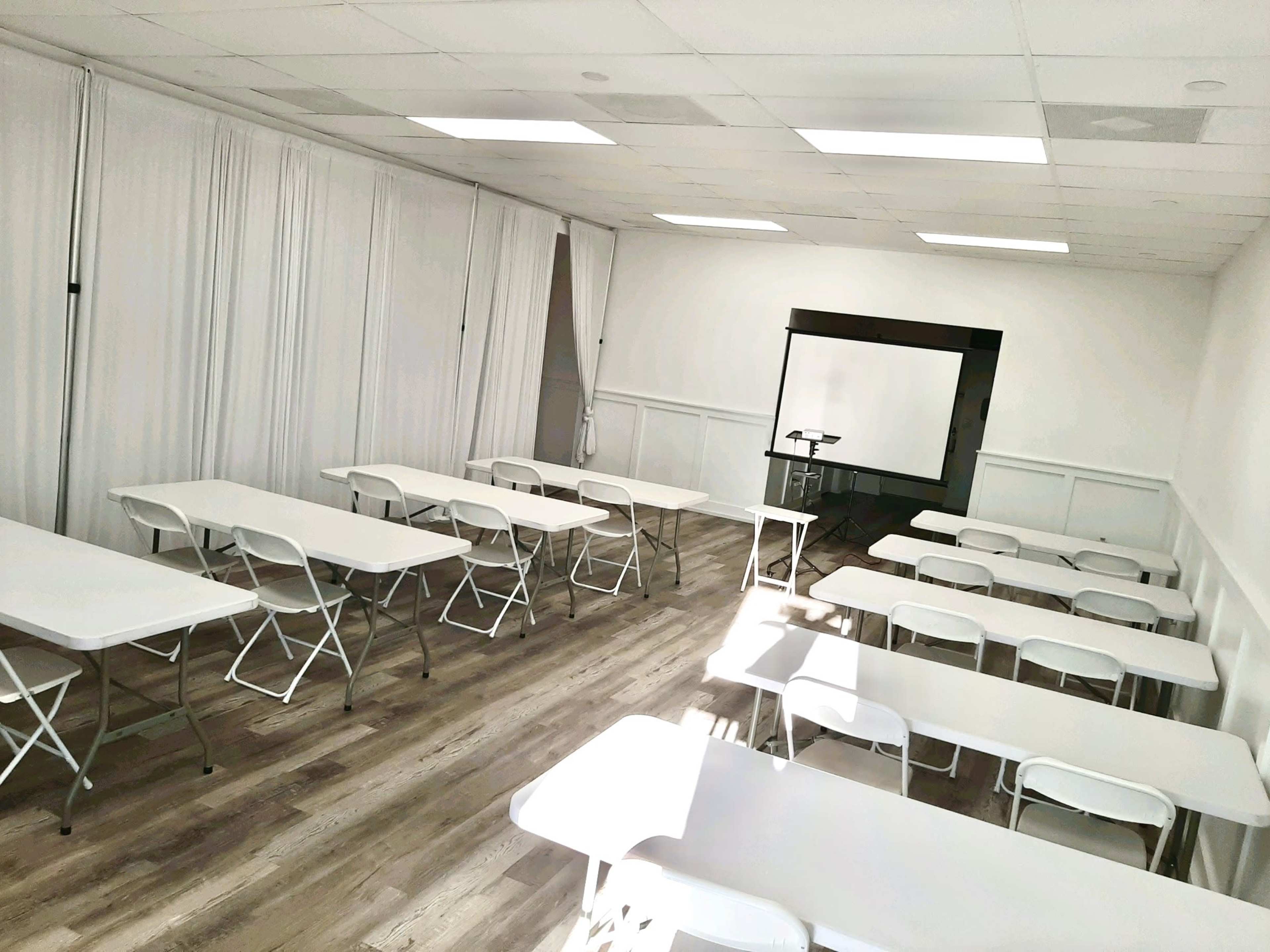 A bright, empty classroom features rows of white folding tables and chairs arranged facing a screen at the front.
