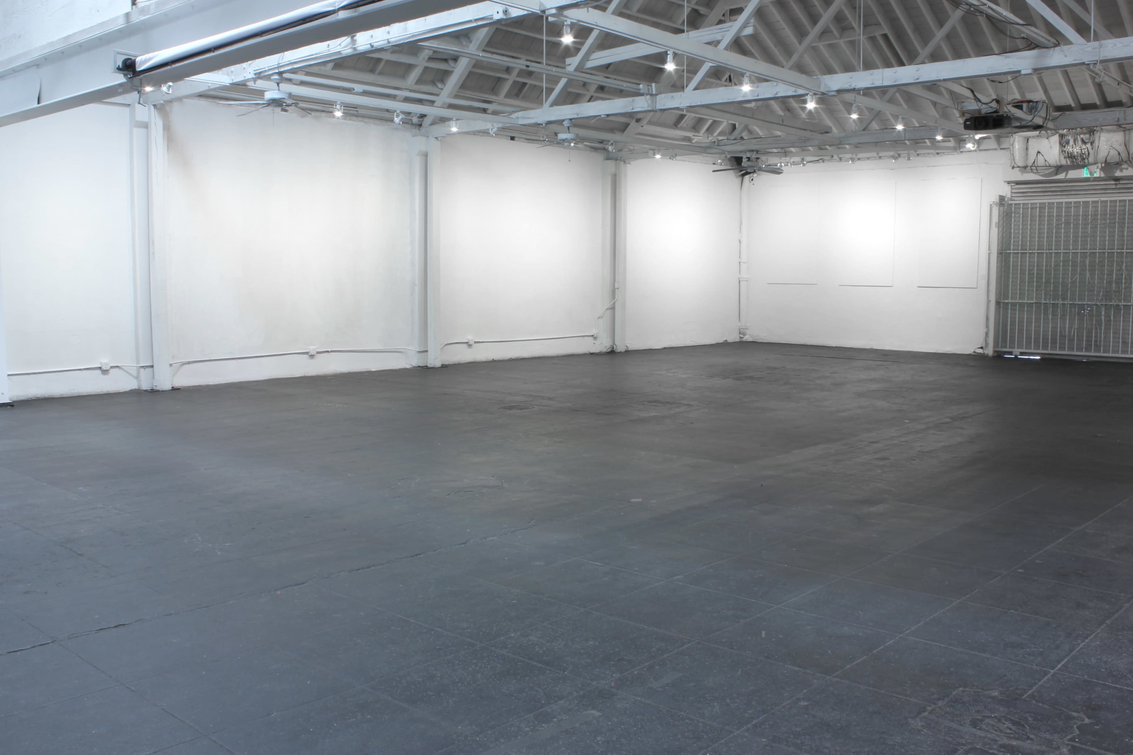 The image shows a large, empty industrial space with black flooring and white walls, featuring exposed beams and overhead lighting.