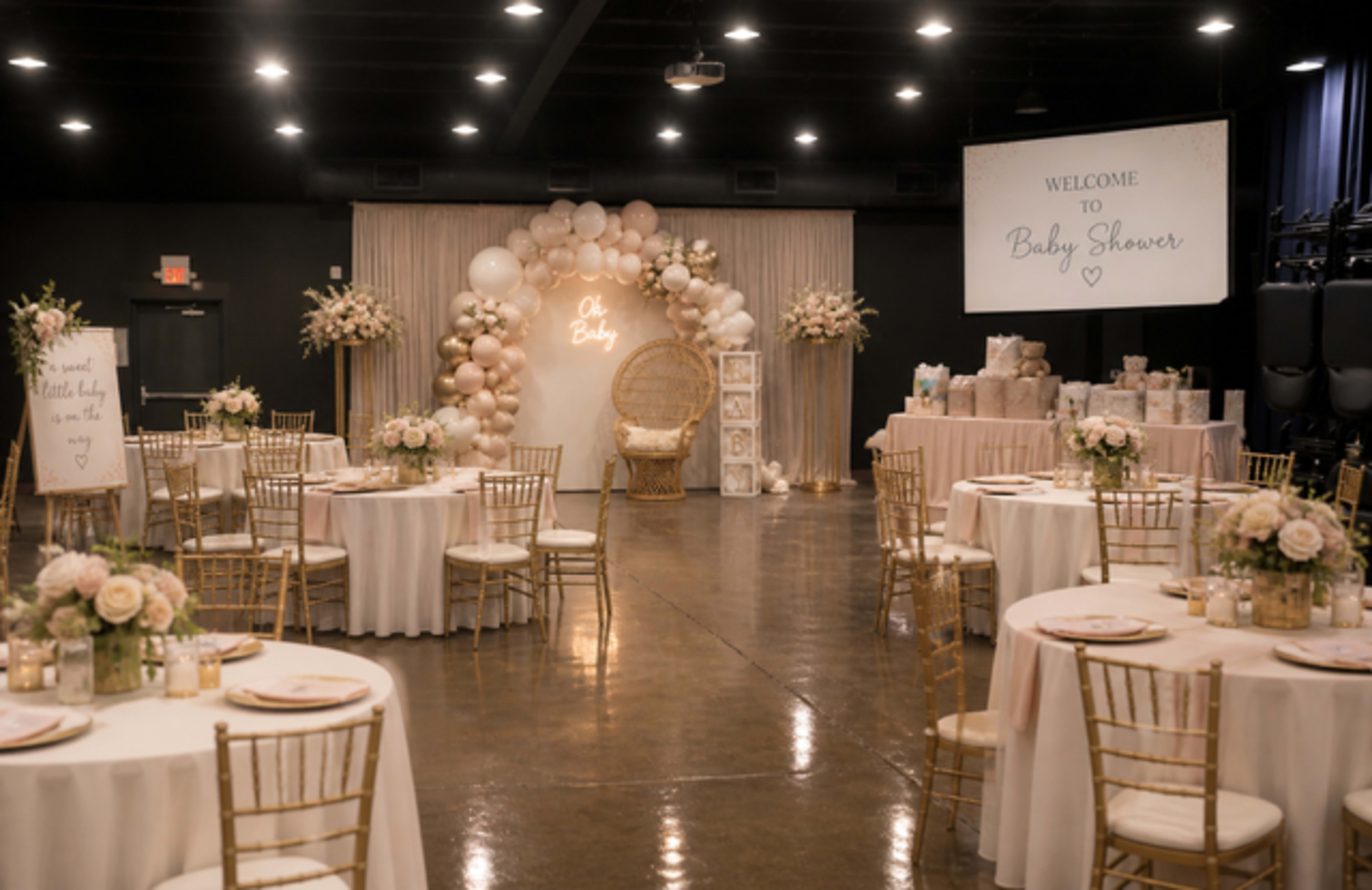 A spacious event hall is set up for a baby shower, featuring elegantly arranged tables with floral centerpieces, a large backdrop of balloons, and a decorative welcome sign.