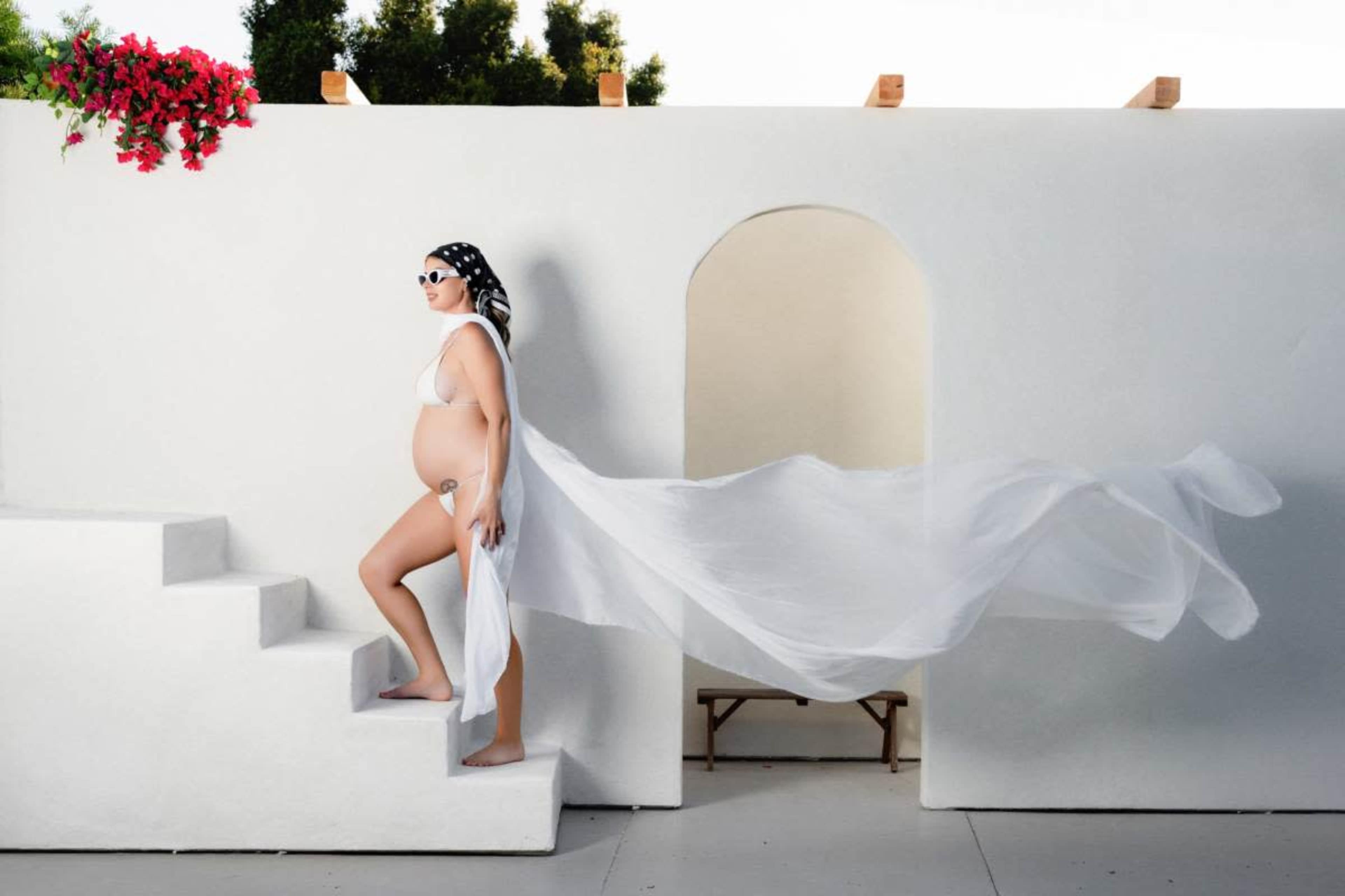 A pregnant woman wearing a white dress ascends a staircase, with a flowing fabric trailing behind her, against a white wall and greenery in the background.