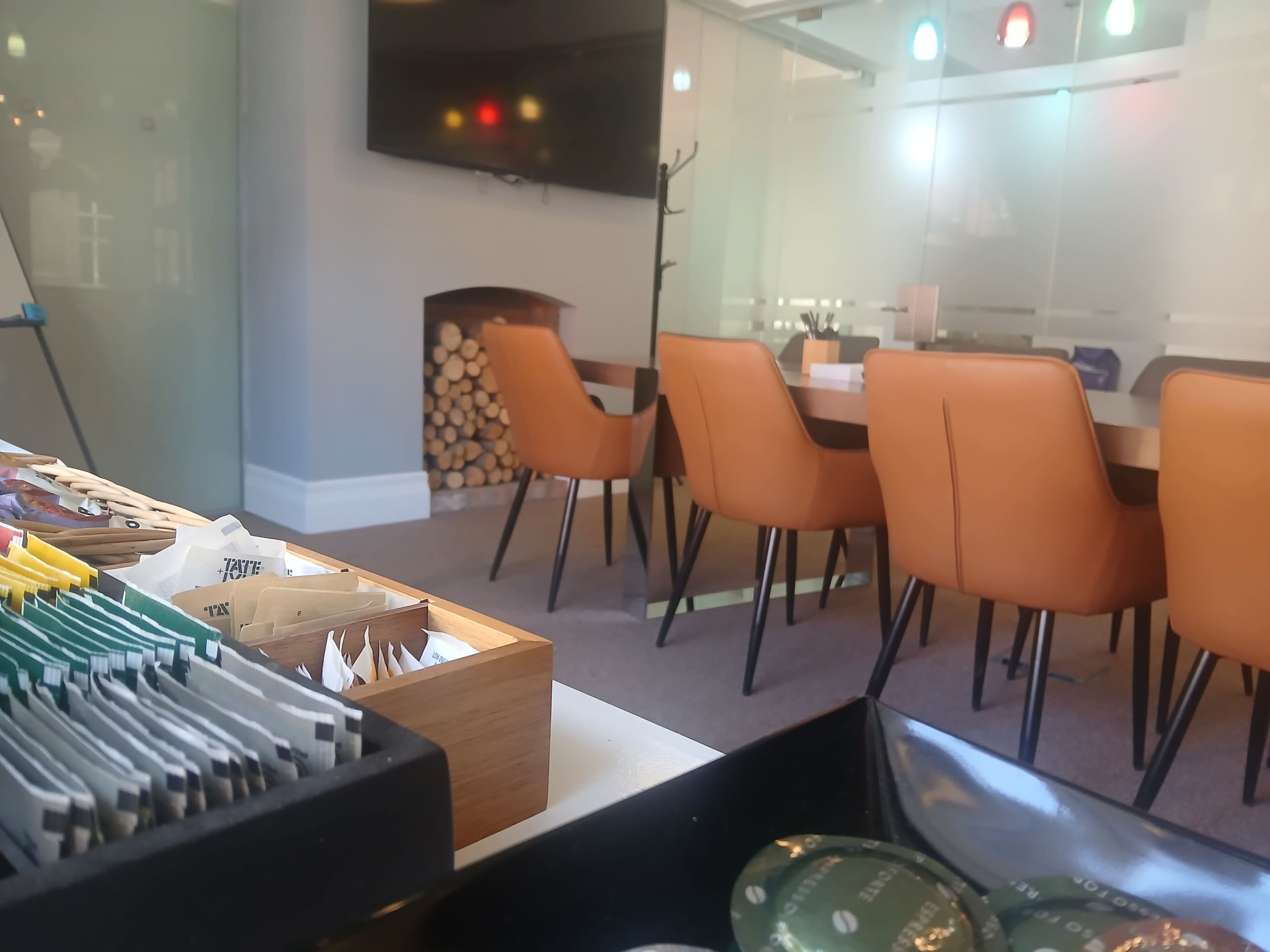 The image shows a modern meeting room with a wooden table surrounded by orange chairs, a television on the wall, and a stack of firewood in the corner.