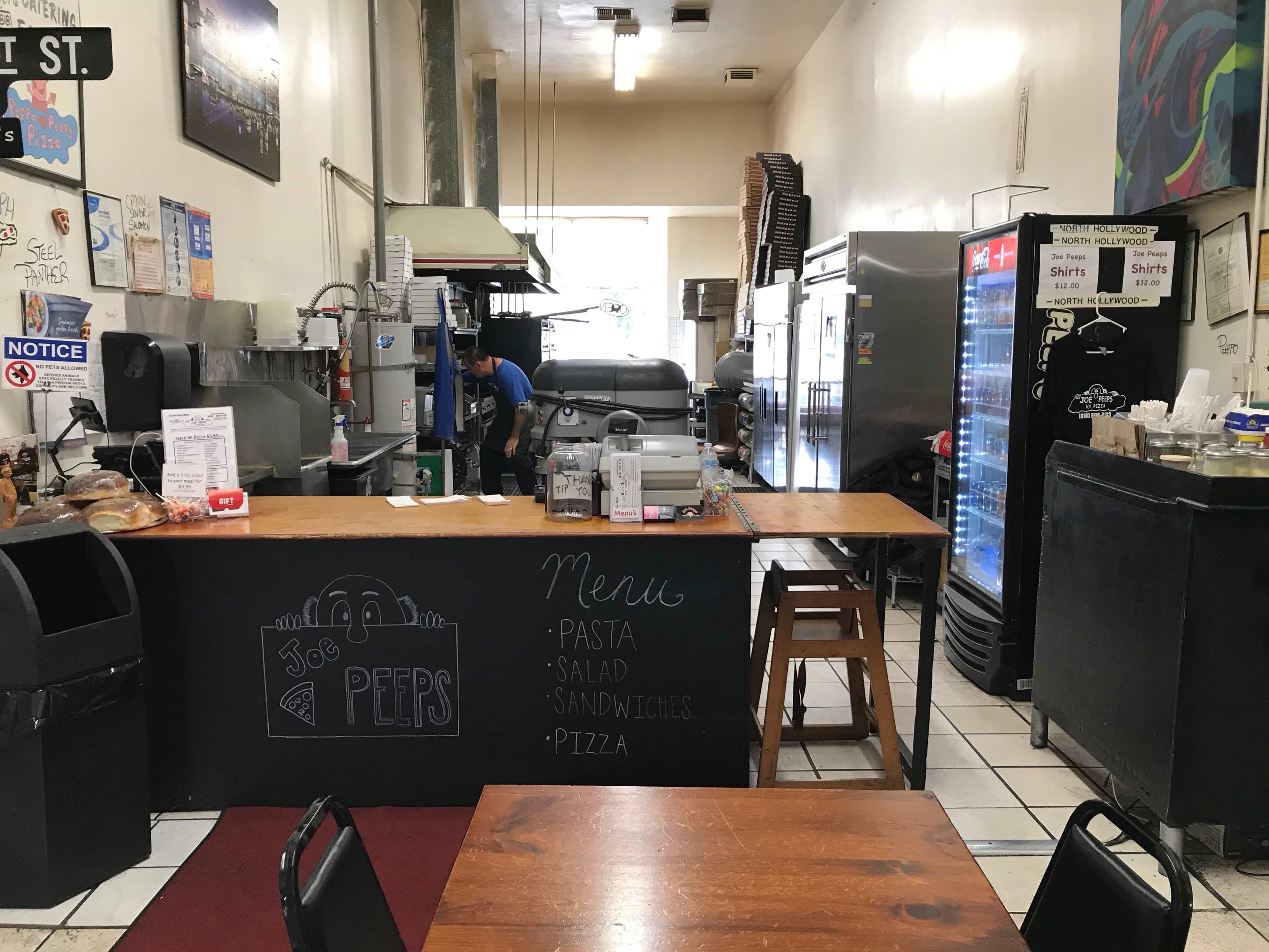 The image shows a small restaurant kitchen with a counter displaying a menu that includes pasta, salad, sandwiches, and pizza.