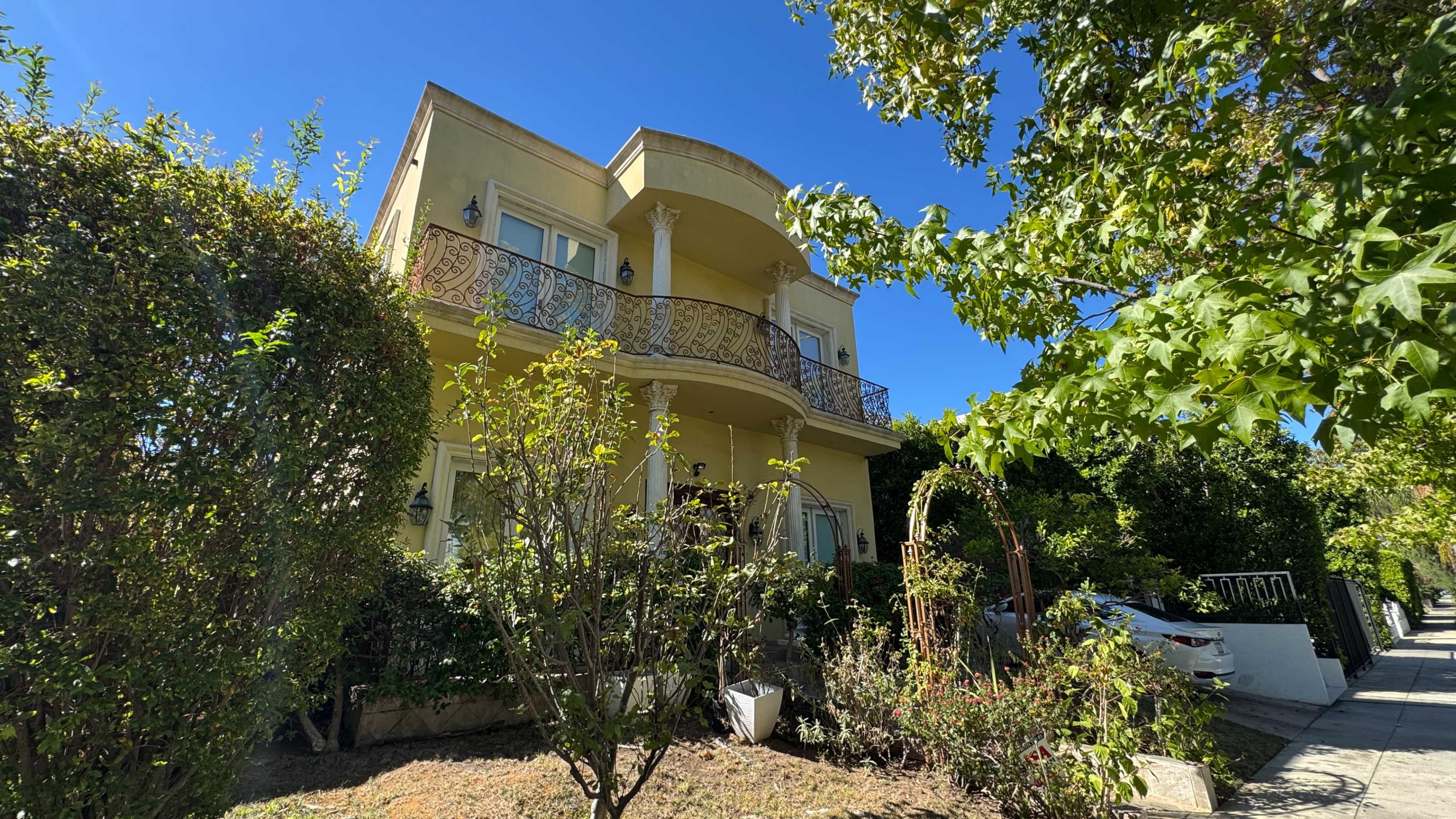 The image shows a two-story yellow house with a balcony and surrounded by lush greenery and shrubs.