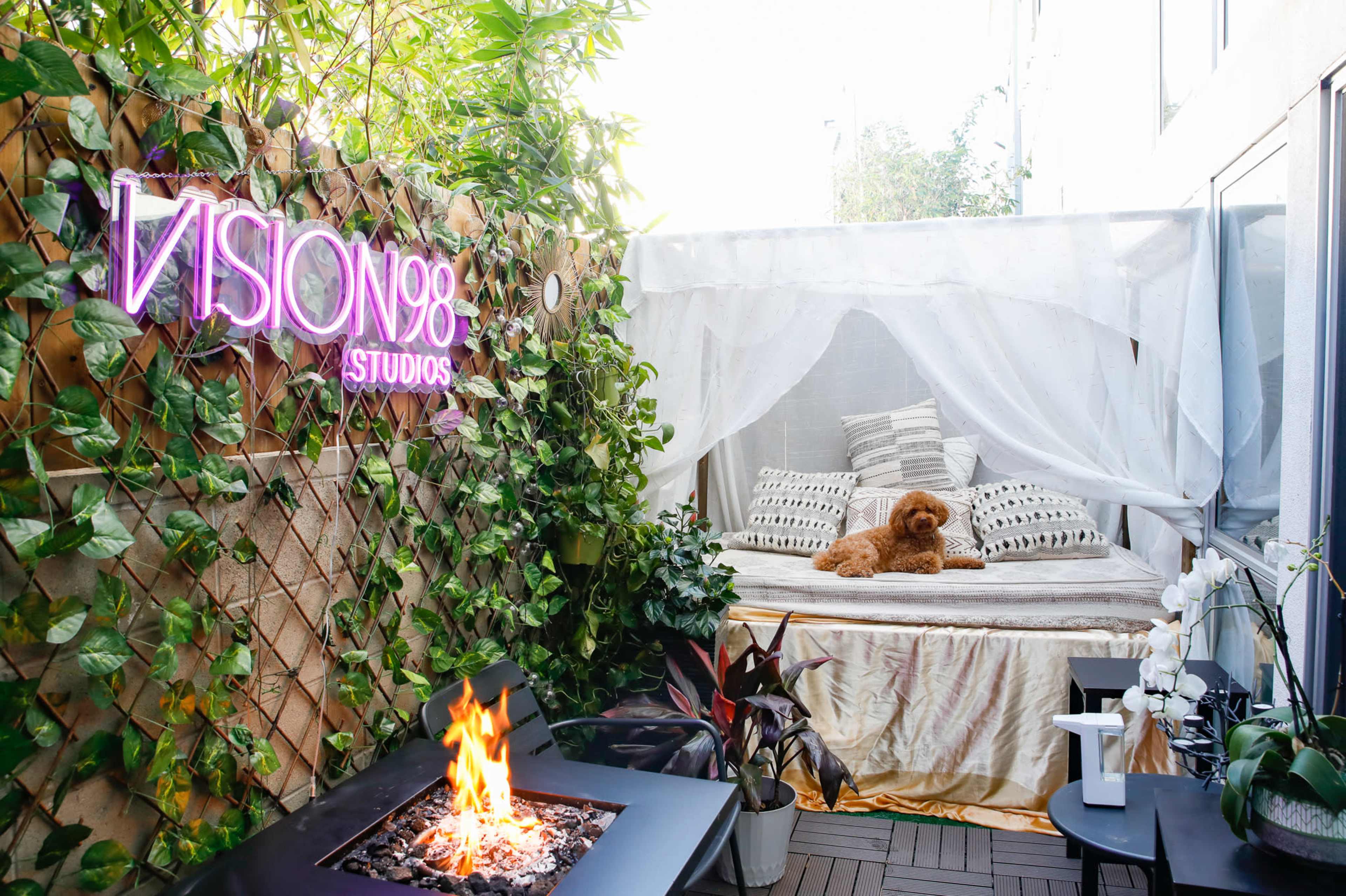 A small outdoor space features a cozy bed draped with sheer fabric, surrounded by greenery and a sleek fire pit.