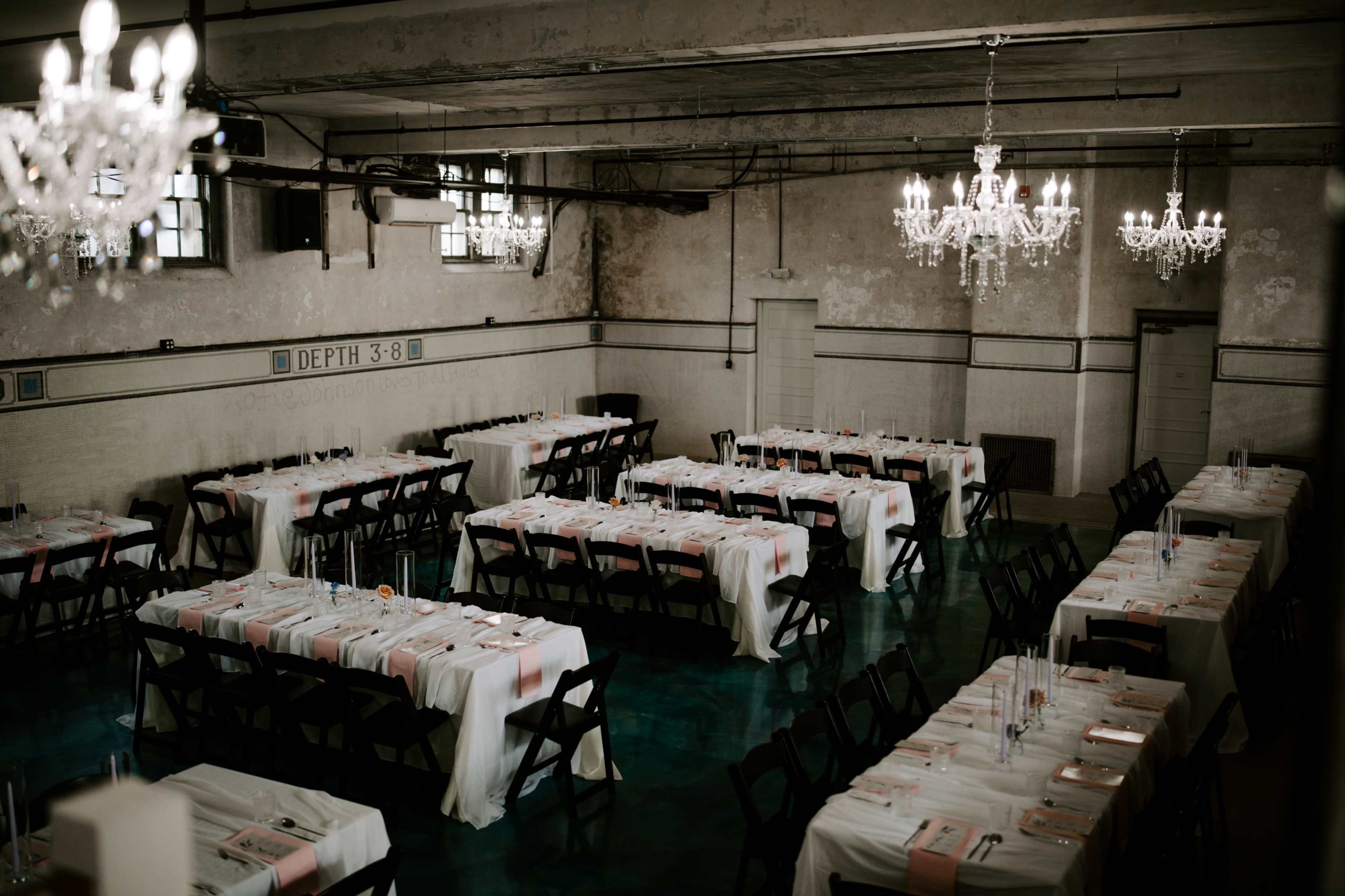 The image shows a large, elegantly arranged dining hall with multiple tables set for a formal event, featuring white tablecloths and pink accent details.