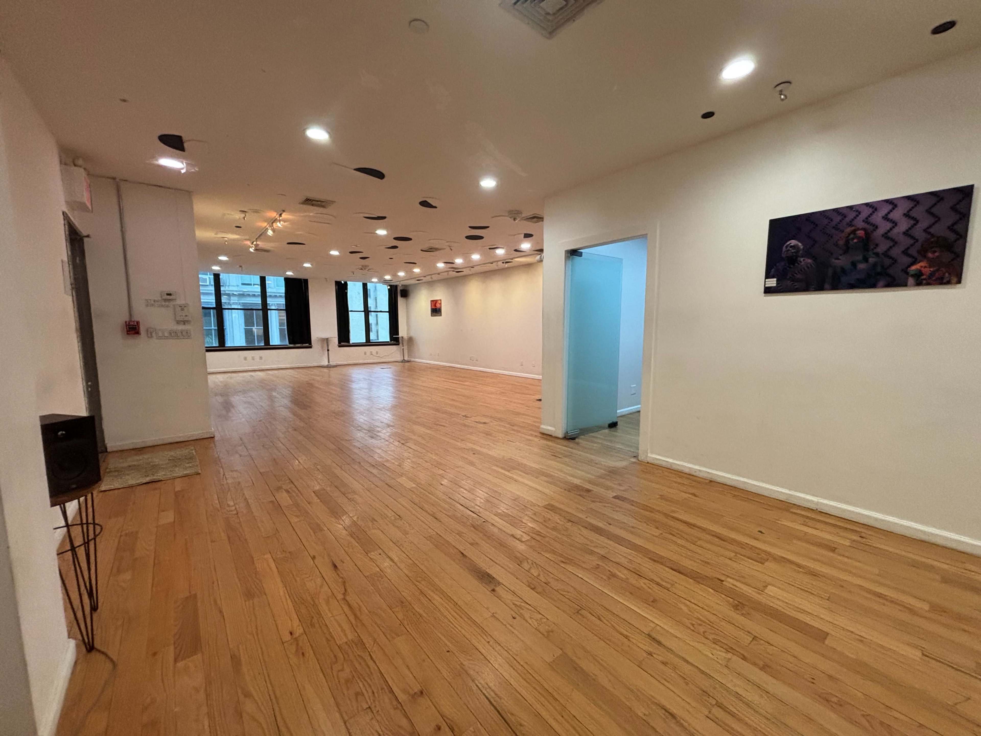 The image shows a spacious, empty room with hardwood flooring, large windows, and white walls adorned with a few artworks.