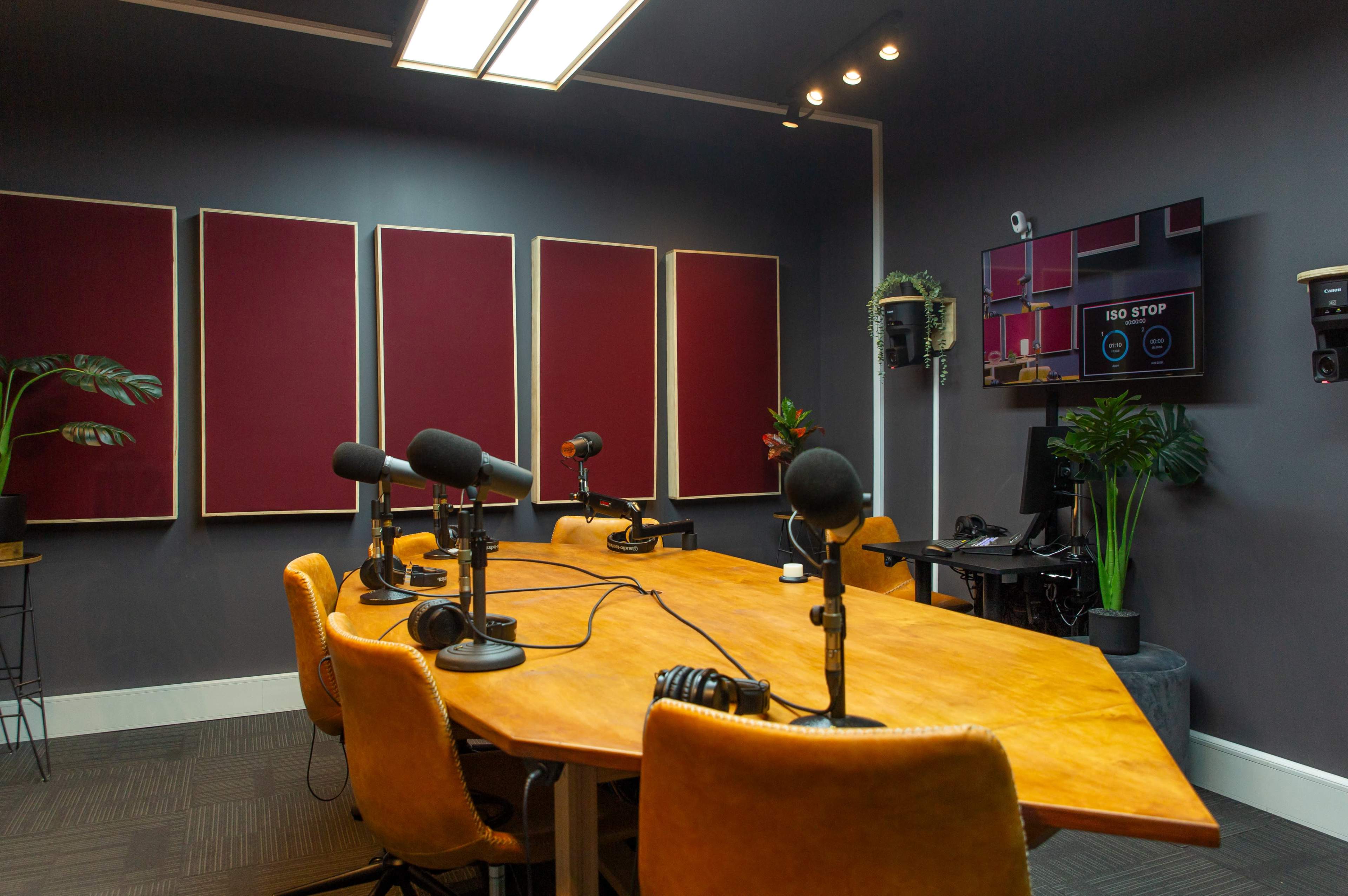 A modern podcast studio features a large wooden table surrounded by several microphones and chairs, with soundproofing panels on the walls and a monitor displaying control options.