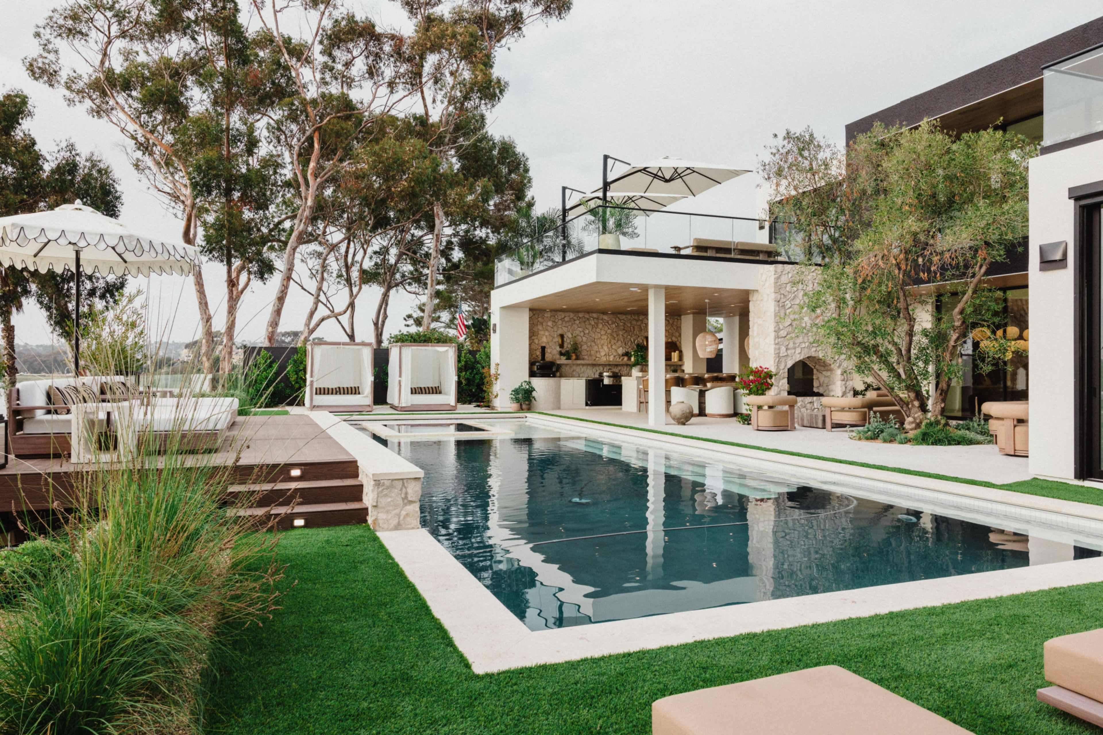The image shows a modern home with a swimming pool surrounded by landscaped green grass and seating areas, featuring outdoor furniture and an umbrella.