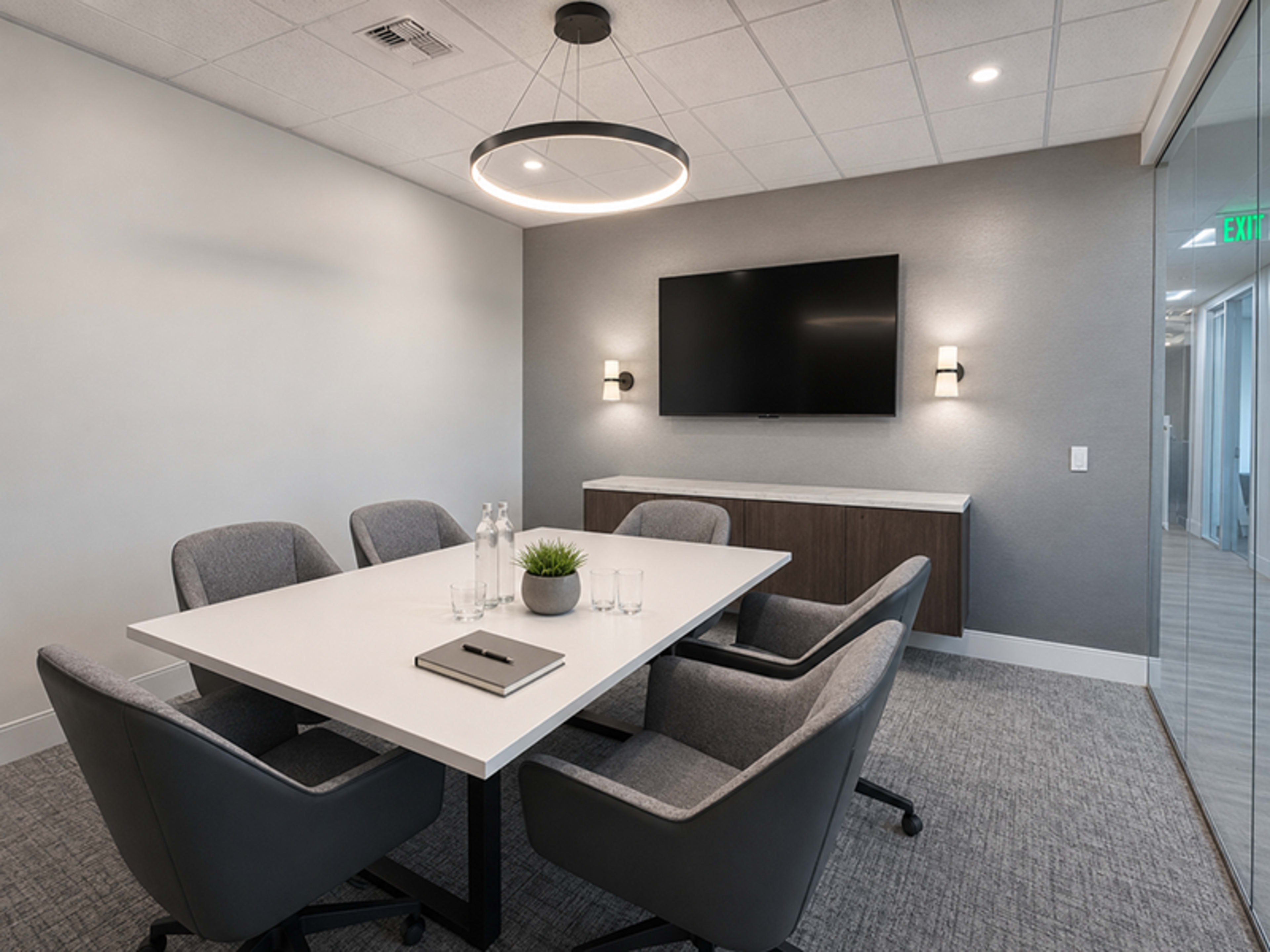 A modern conference room features a large white table surrounded by gray chairs, with a wall-mounted television and a decorative plant centerpiece.