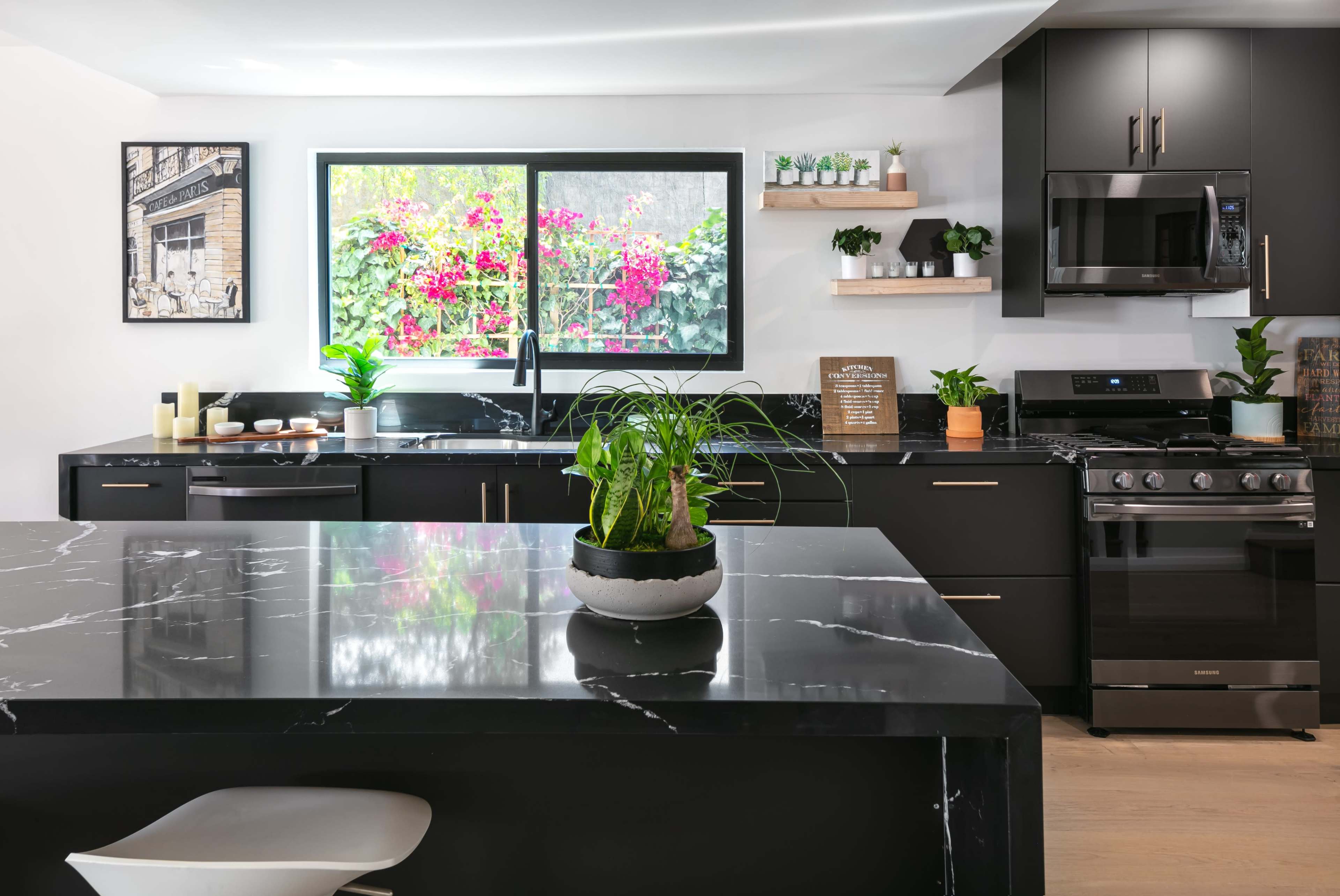 A modern kitchen features dark cabinetry, a black marble countertop with a potted plant, and a window displaying vibrant flowers.