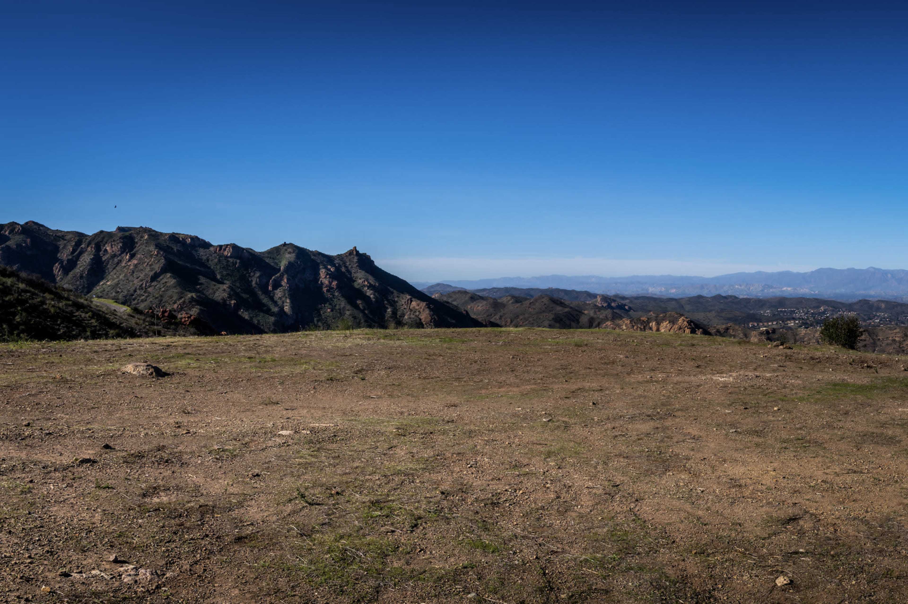 The image shows a clear, flat area overlooking a mountainous landscape under a bright blue sky.
