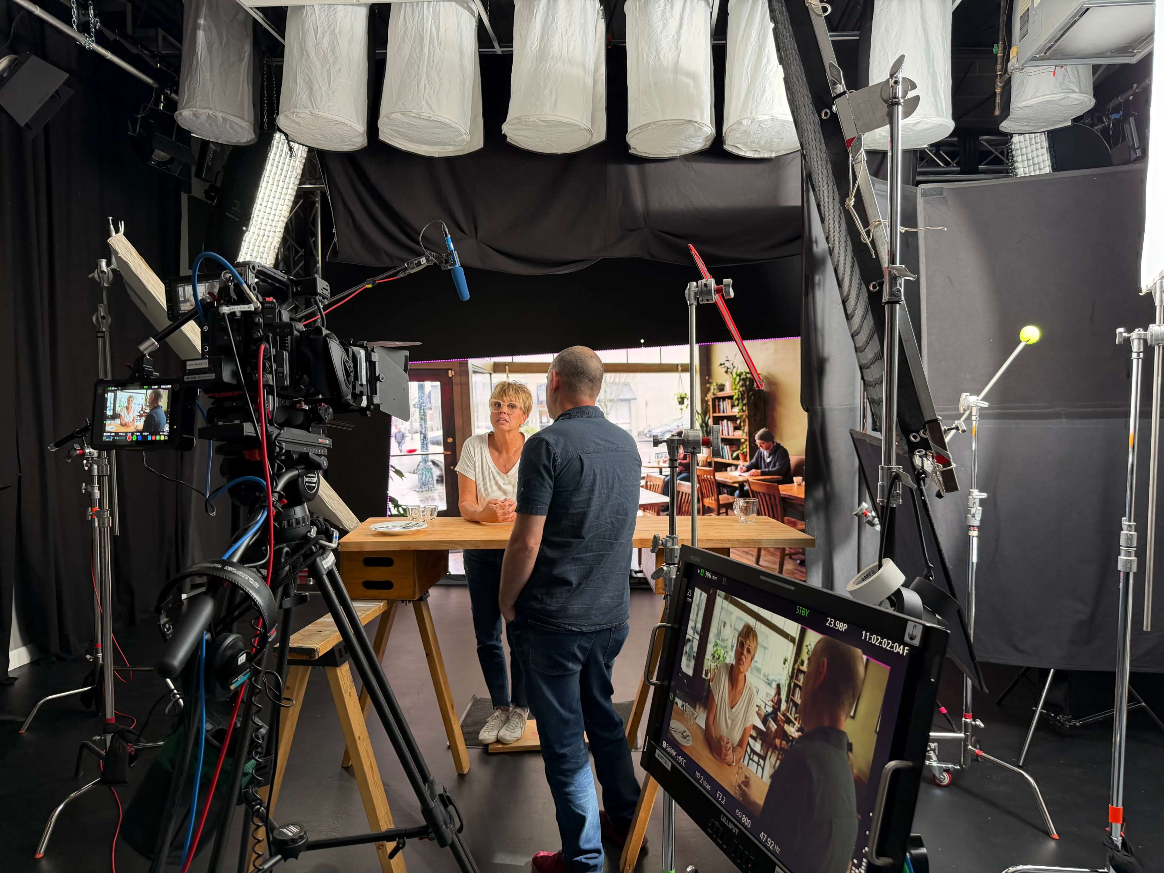 A film crew is set up in a studio with lighting, cameras, and a monitor, where a woman is seated at a table while a man stands nearby.