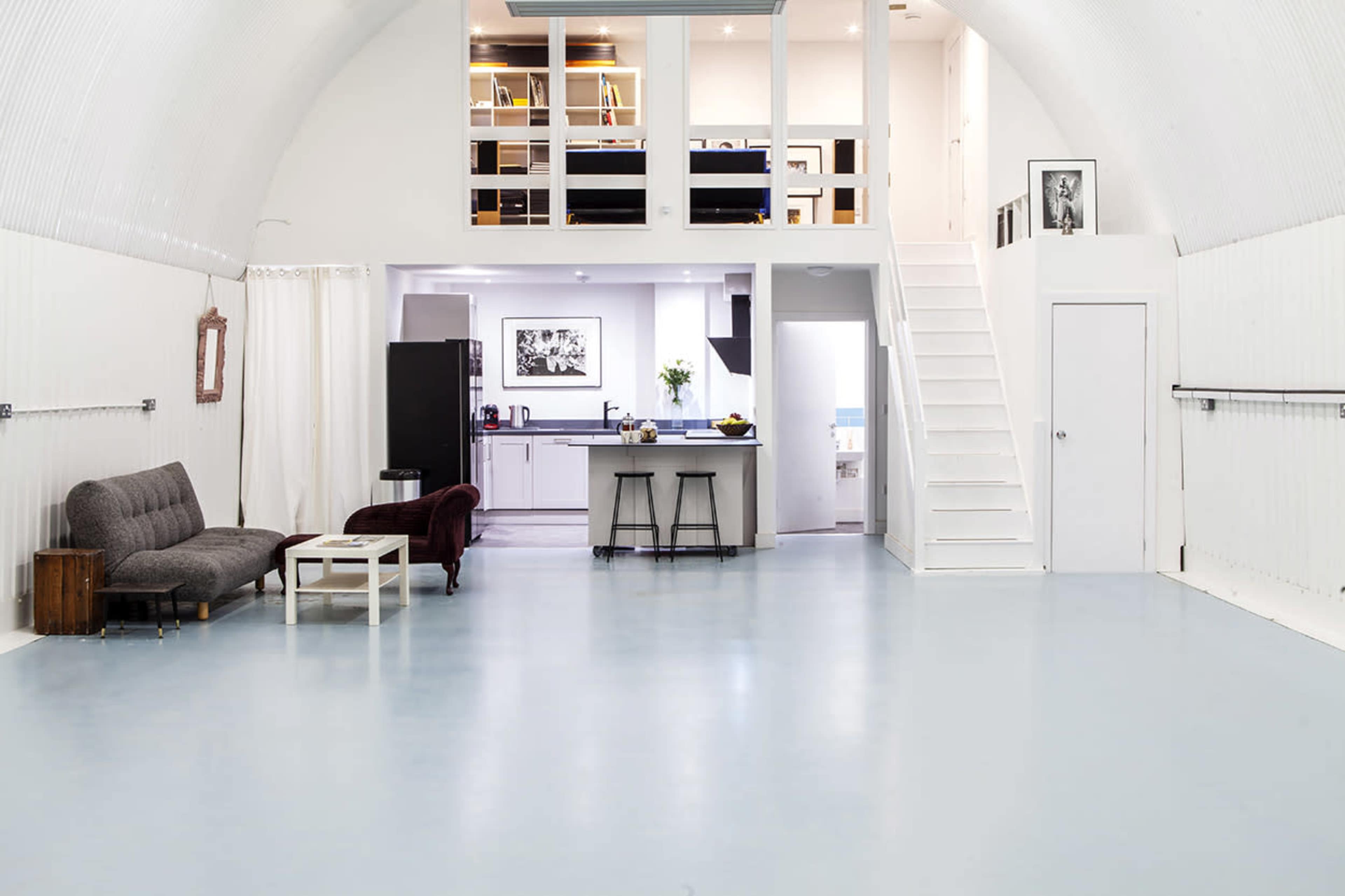 The image shows a spacious, modern room with a high ceiling, featuring a living area with a gray sofa, a burgundy chair, and a small white table, leading to a kitchenette and a staircase.
