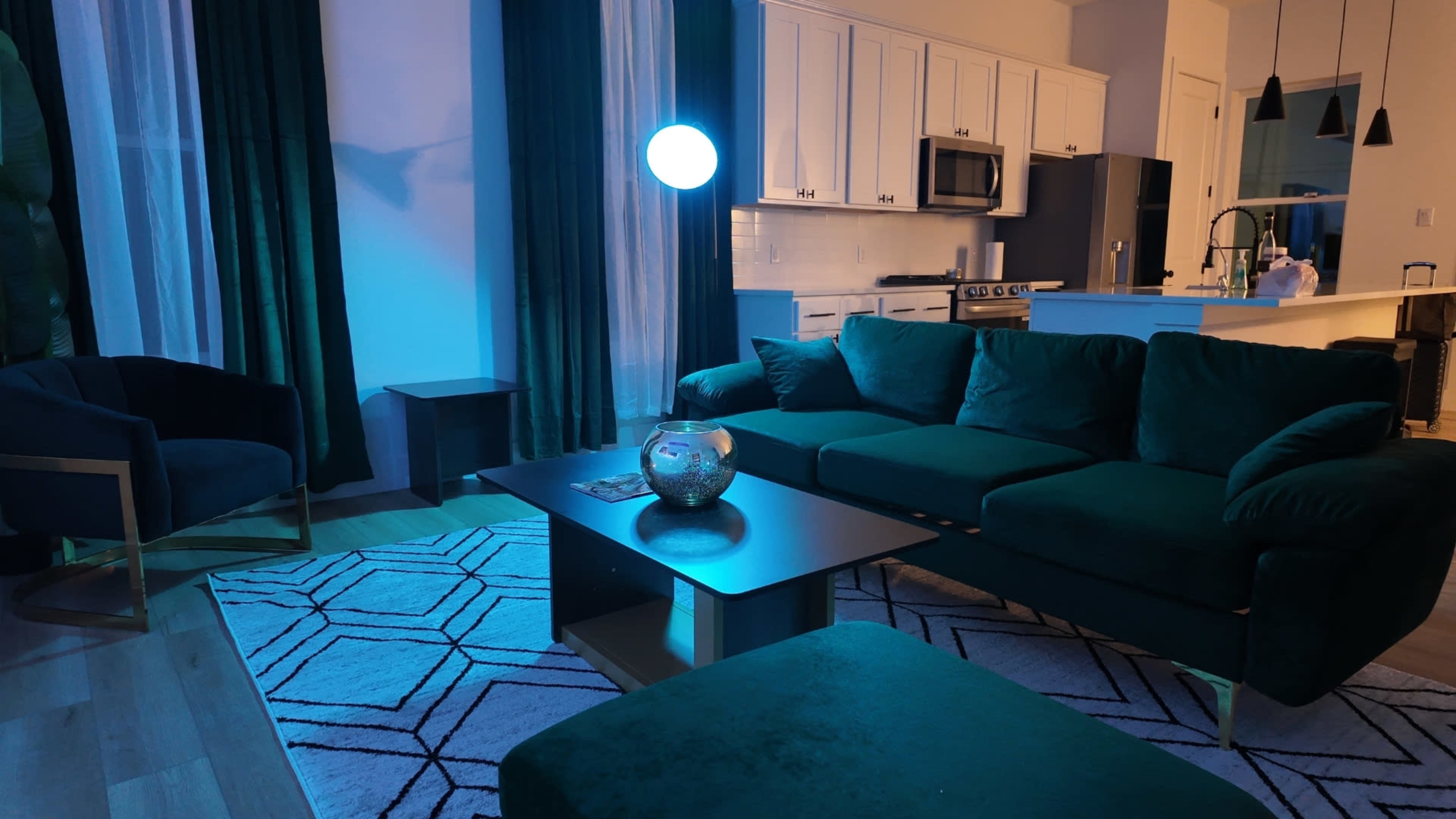 A modern living room features a dark green sofa, a geometric area rug, and a round blue pendant light, with kitchen appliances visible in the background.