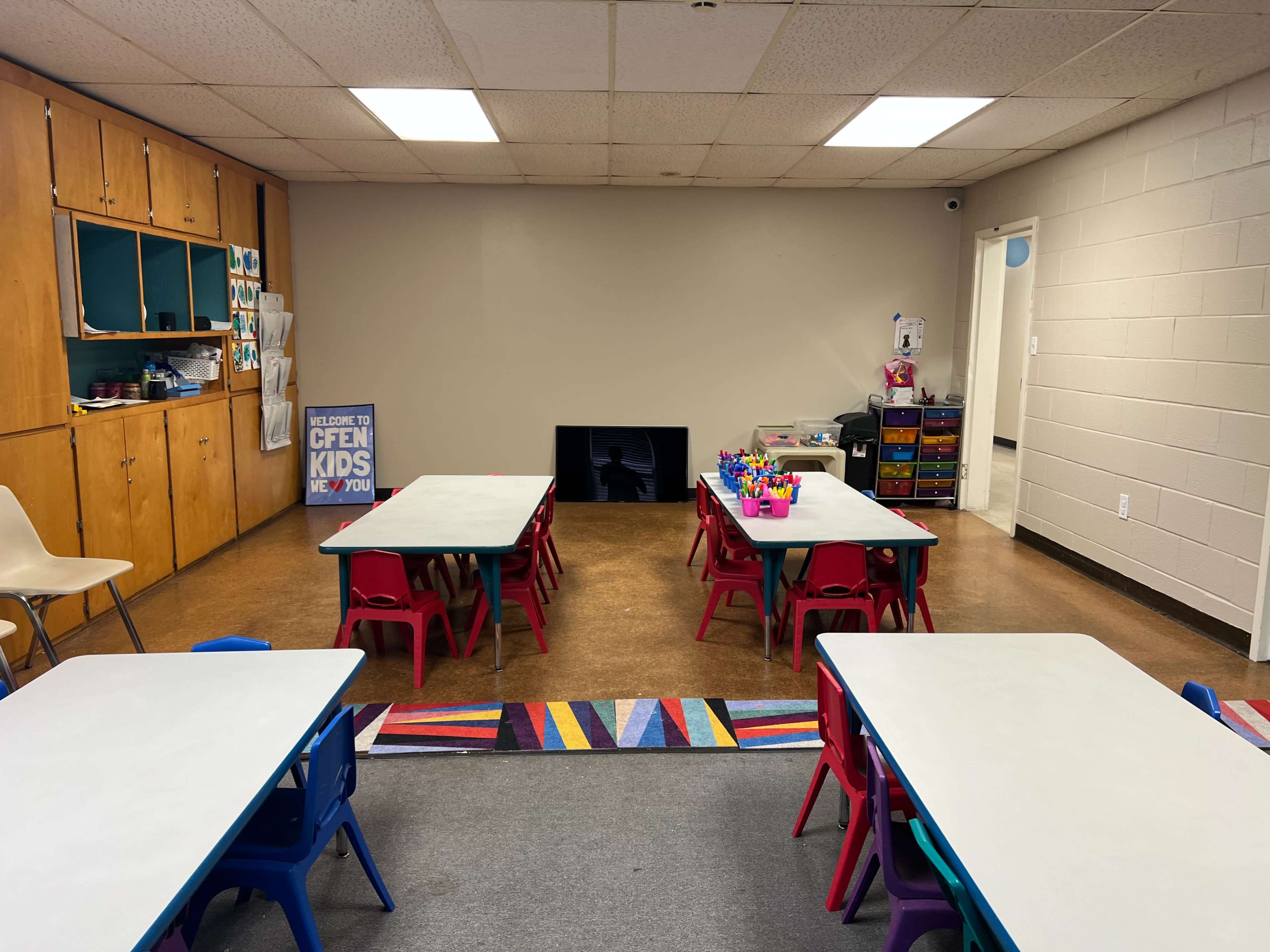A classroom features multiple tables with colorful chairs, organized supplies, and a large empty wall.