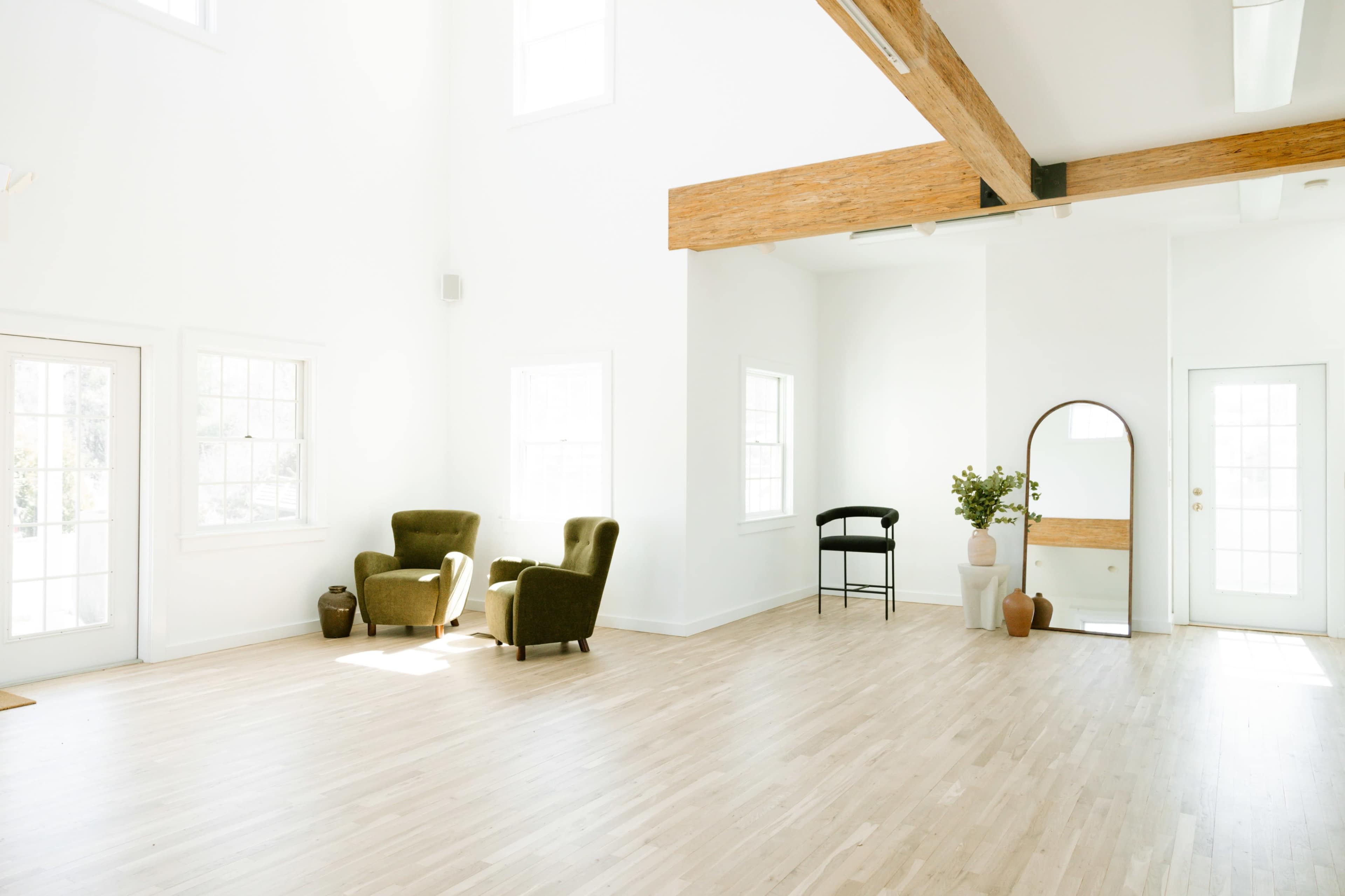 The image showcases a spacious, sunlit interior with light wooden flooring, two green armchairs, and large windows.