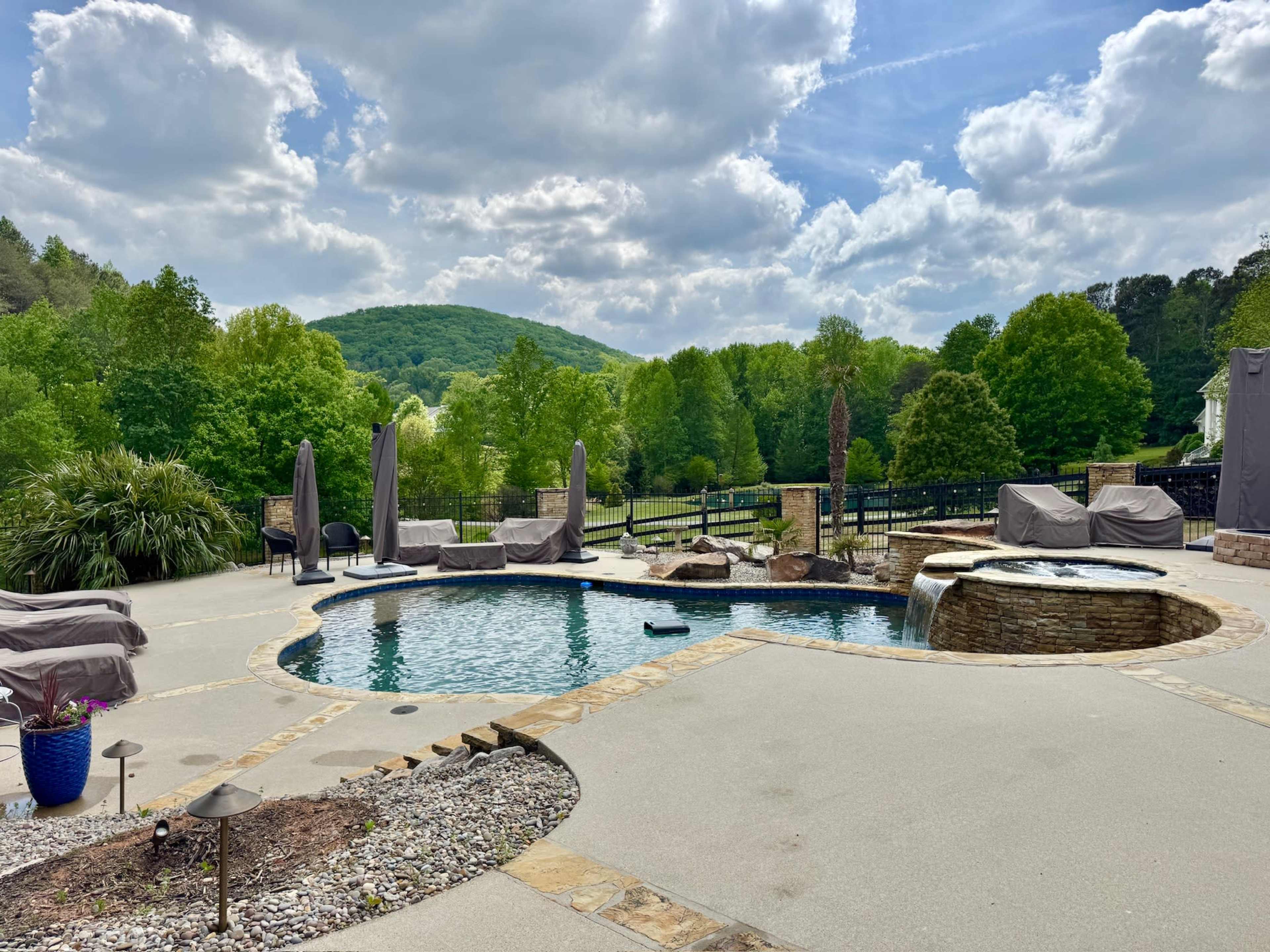 The scene features a landscaped backyard with a swimming pool, a spa area, and umbrellas, set against a backdrop of green hills and a cloudy sky.