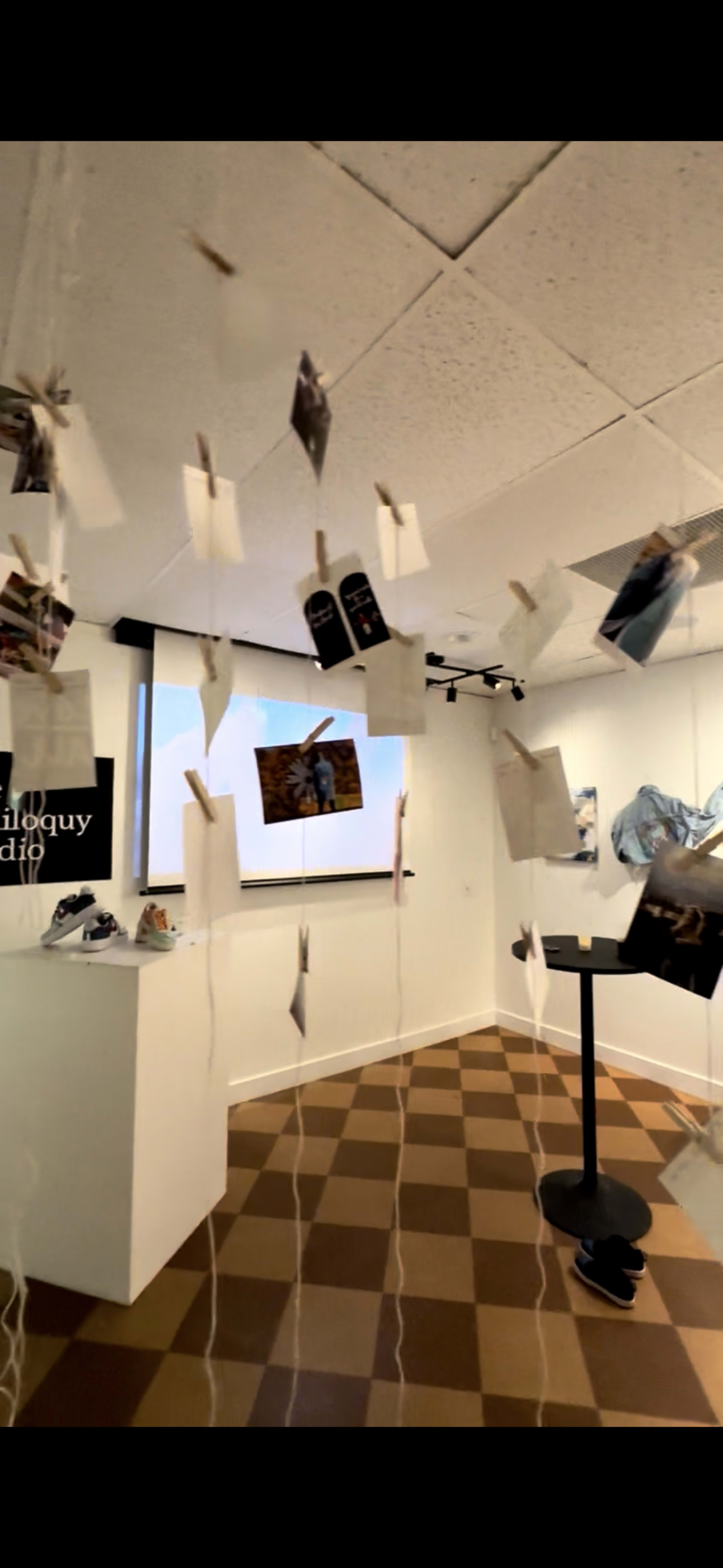 The image shows an art gallery with hanging photographs and display pieces arranged in a room featuring a checkered floor and a presentation screen.