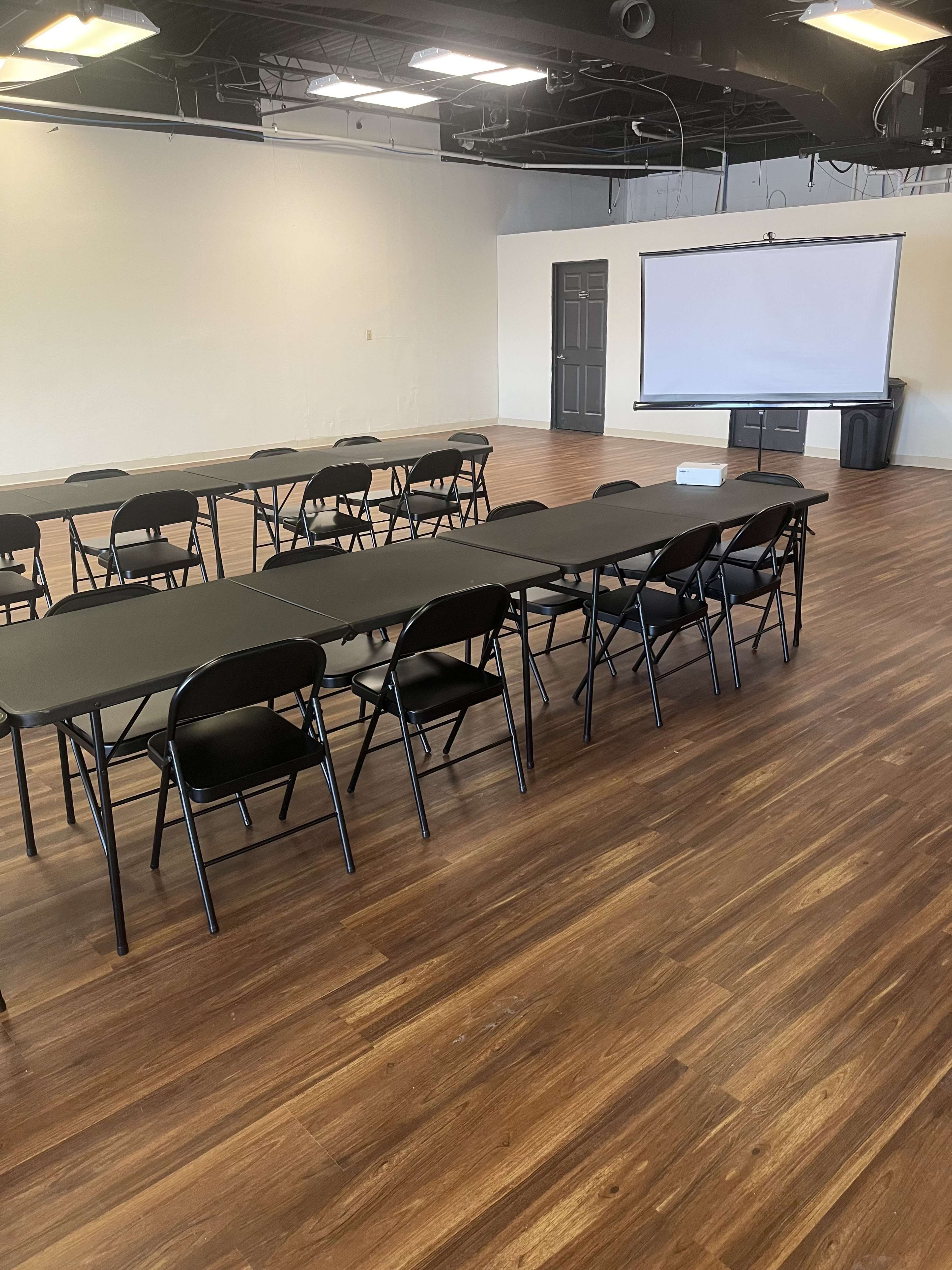 The image shows a spacious room with black rectangular tables and folding chairs arranged in rows, along with a blank projection screen mounted on a wall.
