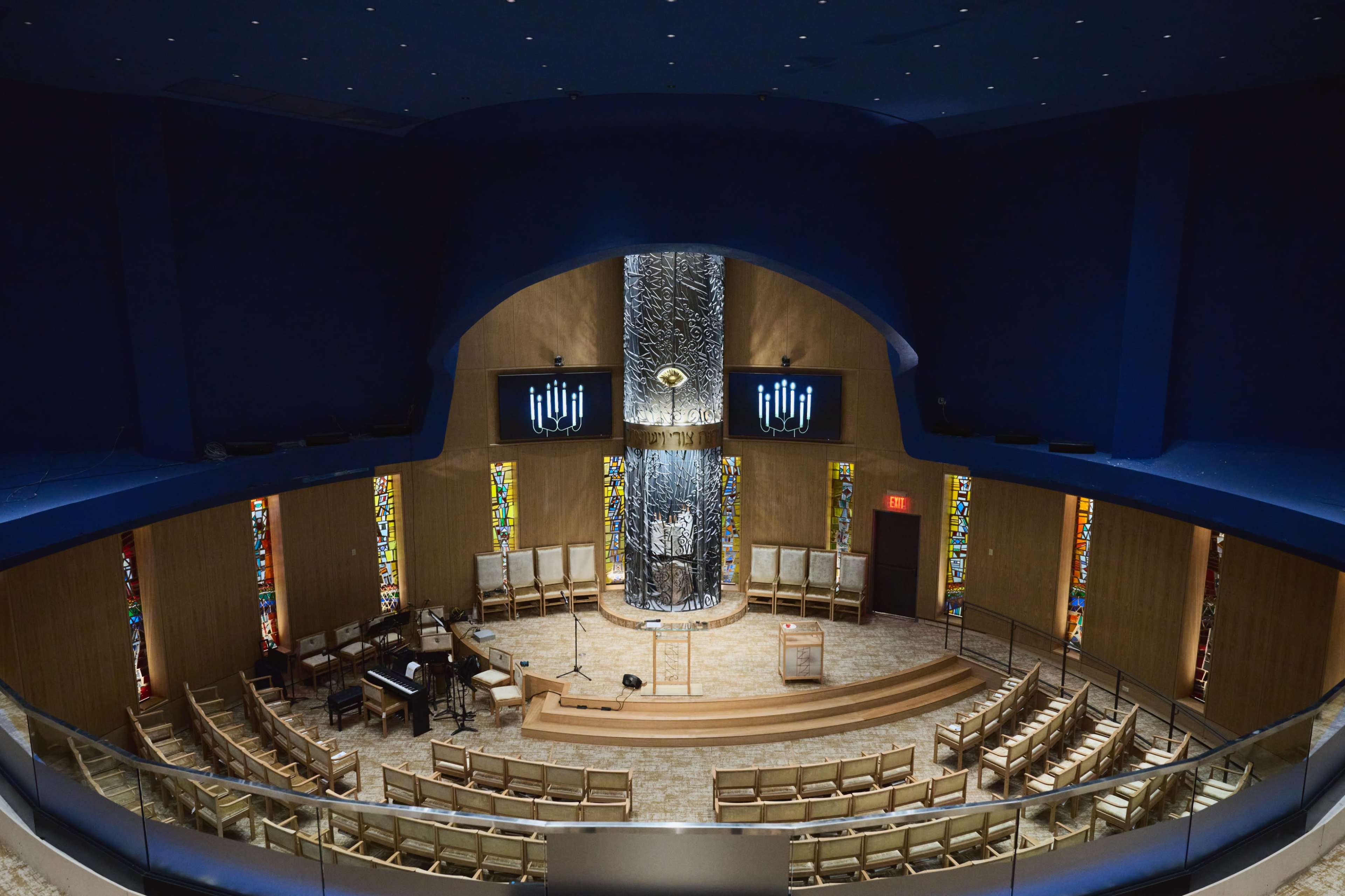 A circular interior of a modern synagogue featuring a central stage flanked by stained glass windows and surrounded by tiered seating.