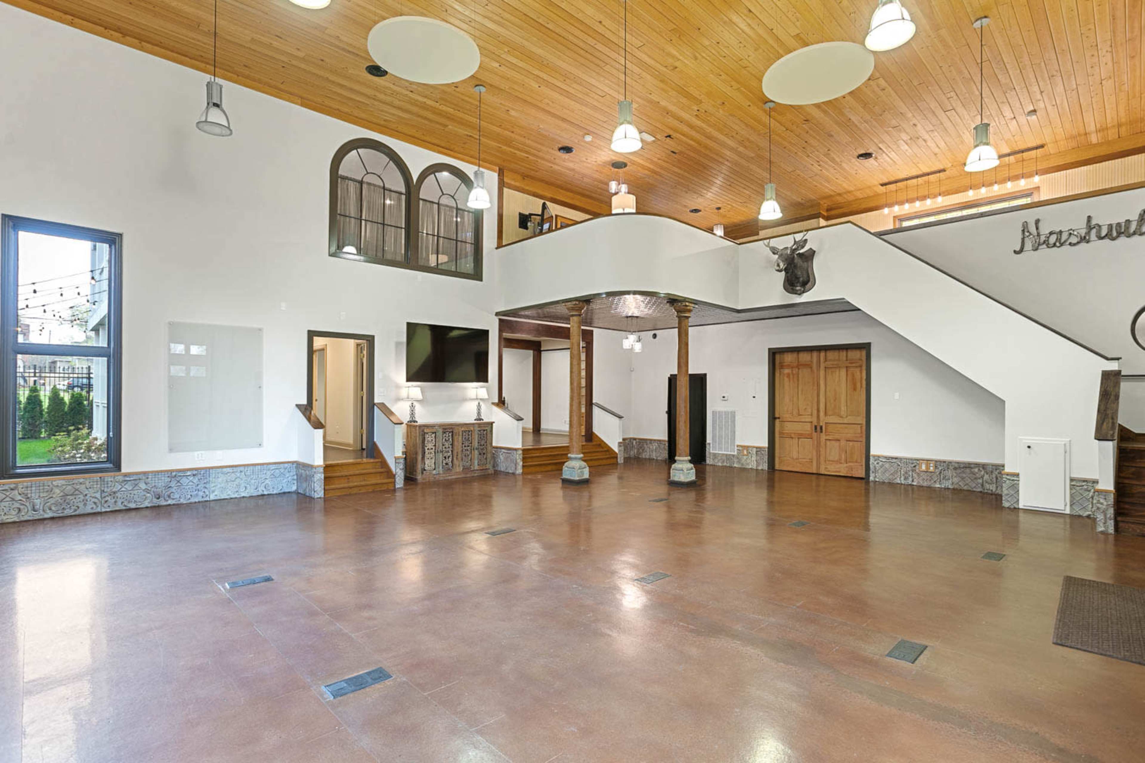The image shows a spacious interior with a wooden ceiling, polished concrete floor, and large windows, featuring a staircase and decorative elements.