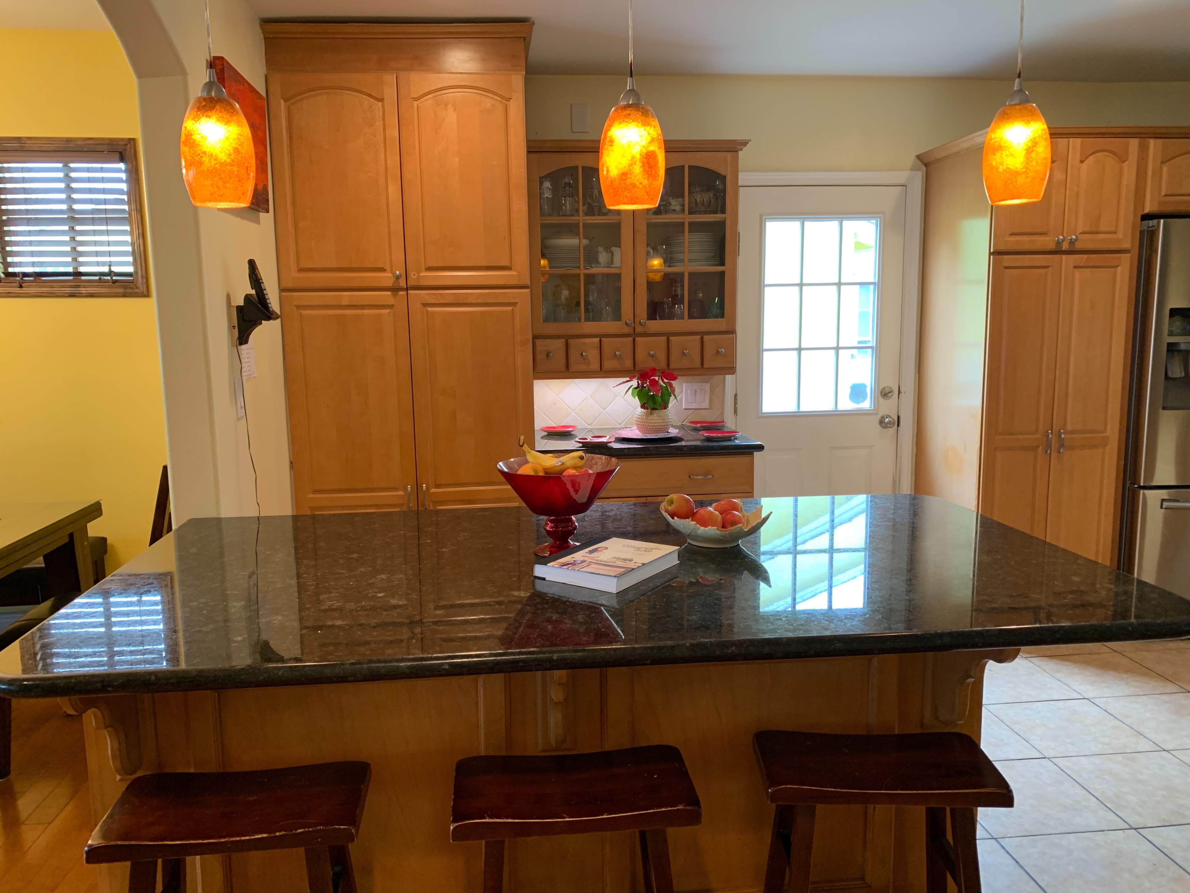 A kitchen features wooden cabinetry, a granite island with bar stools, and pendant lights hanging above.