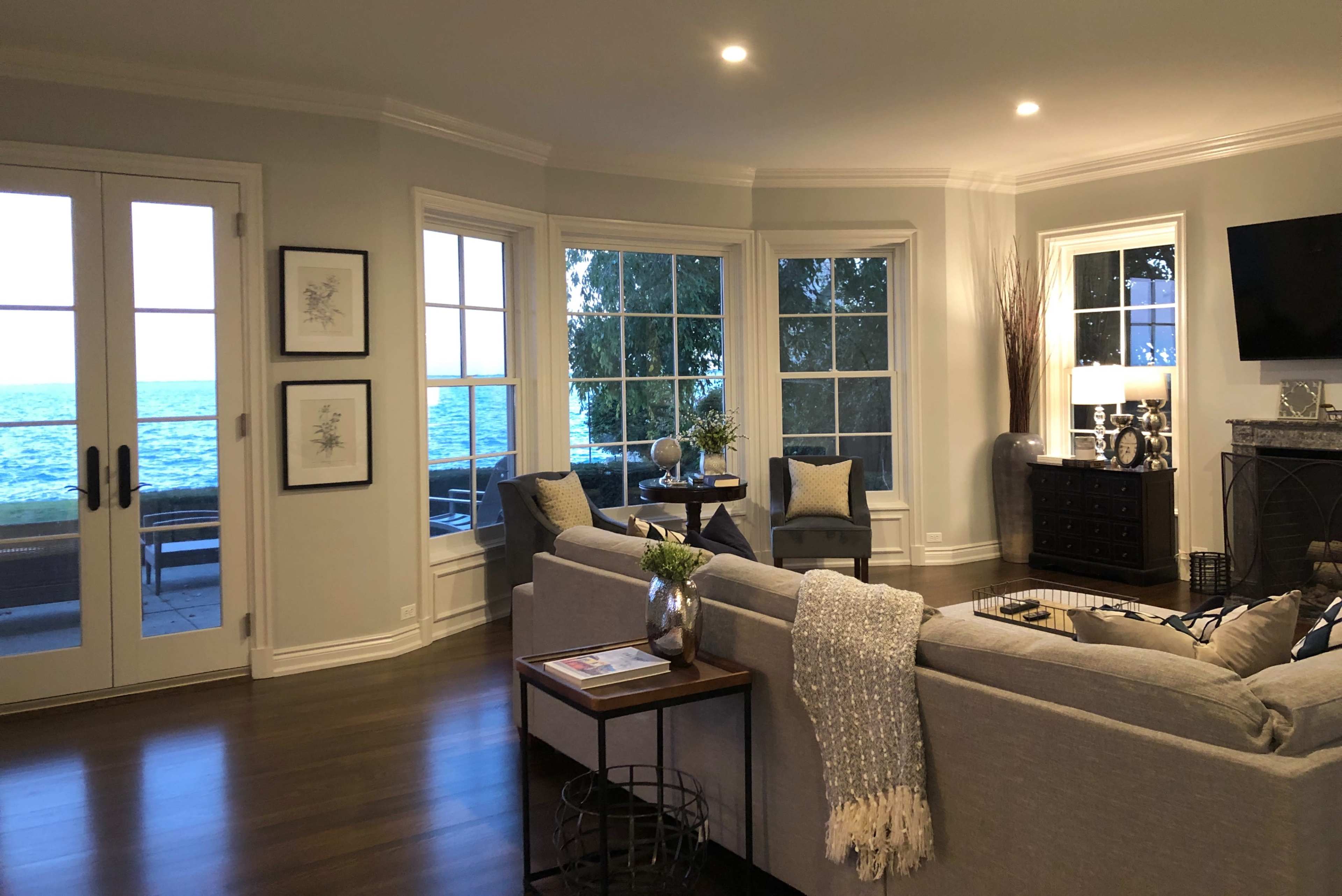 The living room features large windows overlooking a body of water, with a sofa, decorative pillows, a coffee table, and a media console.