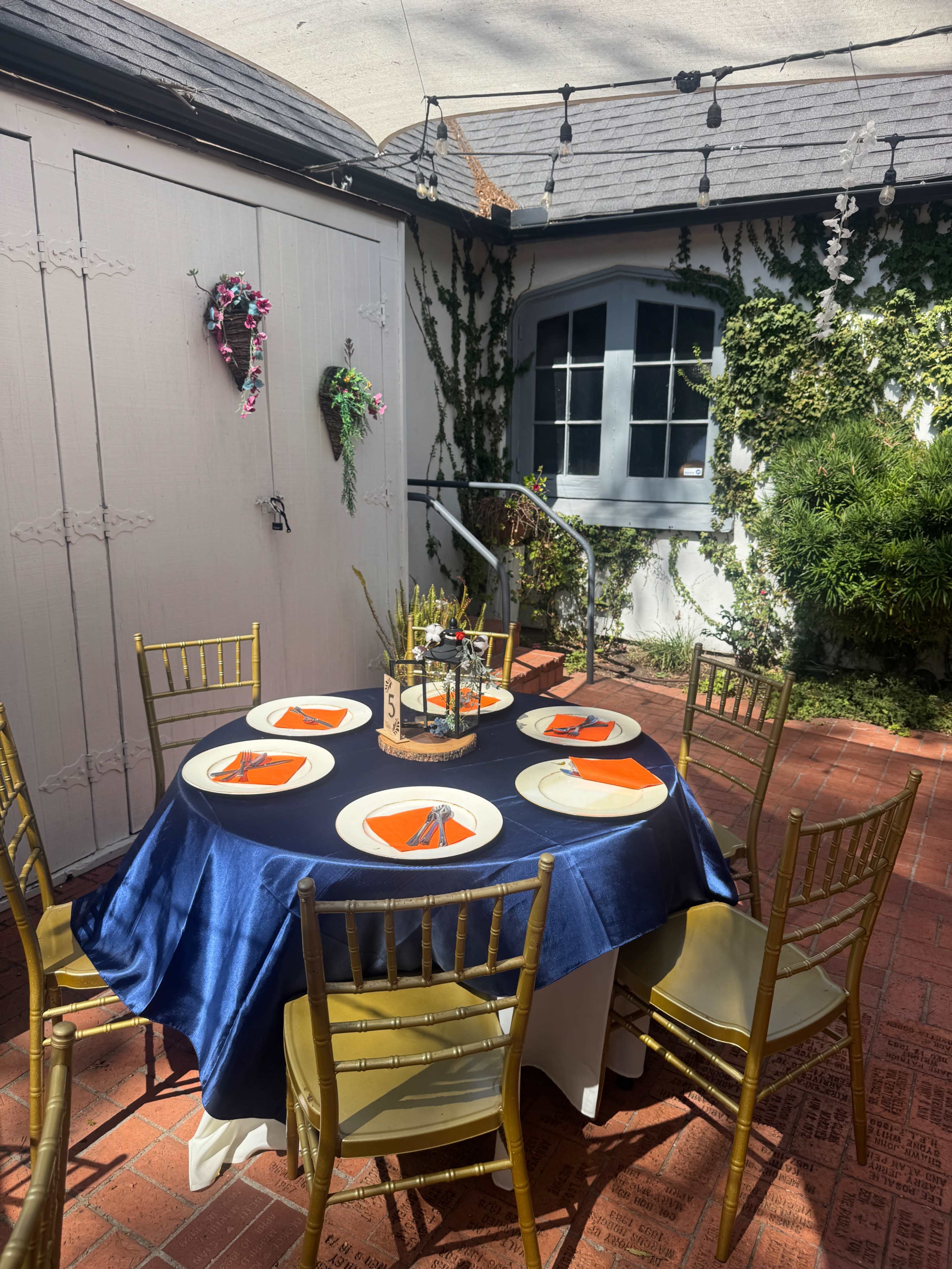 A round table with a navy blue tablecloth is set with orange plates and utensils, surrounded by golden chairs in a courtyard adorned with greenery and decorative elements.