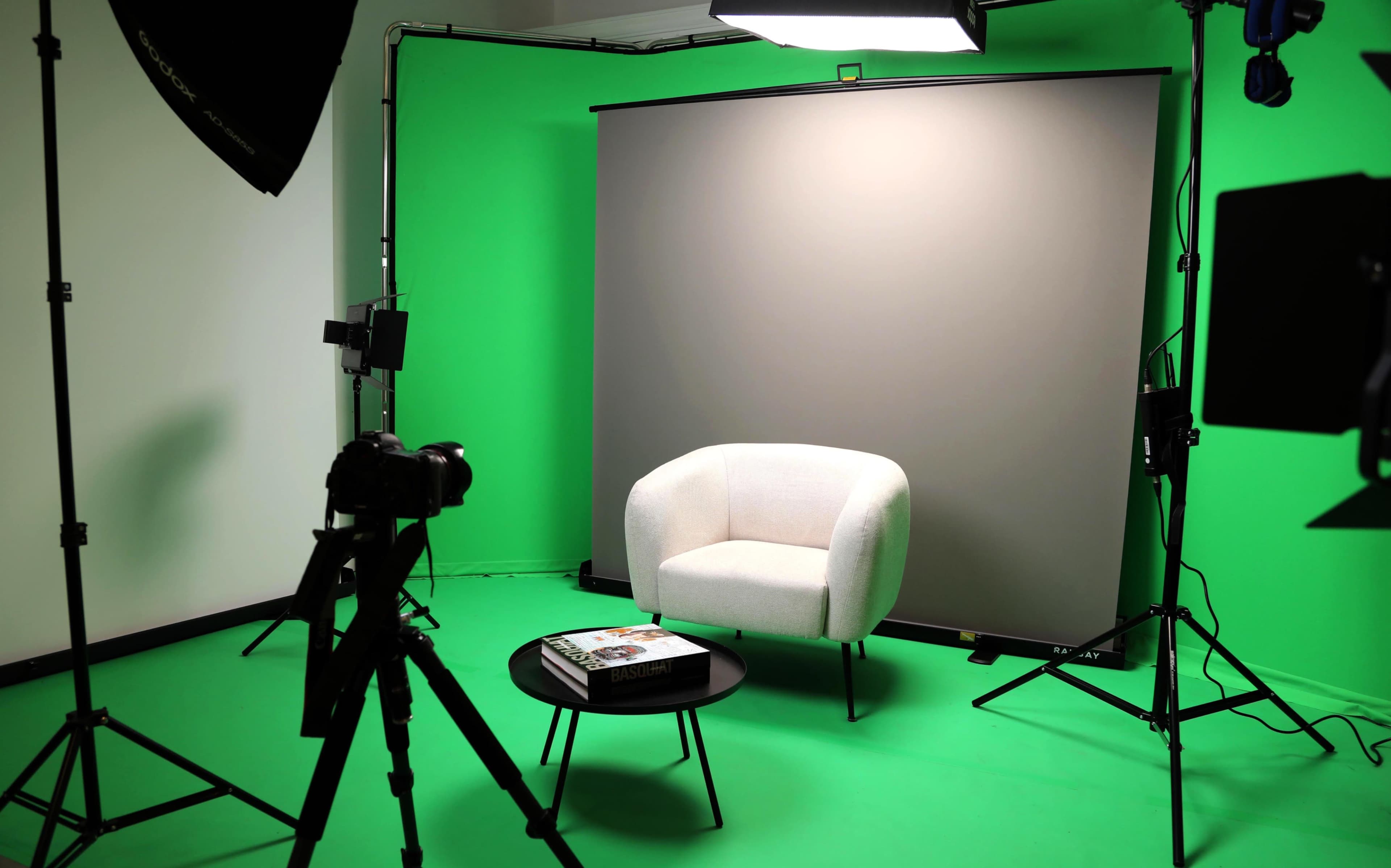 The image shows a photo studio setup with a green backdrop, a light, a camera on a tripod, a small black table, and a white armchair.