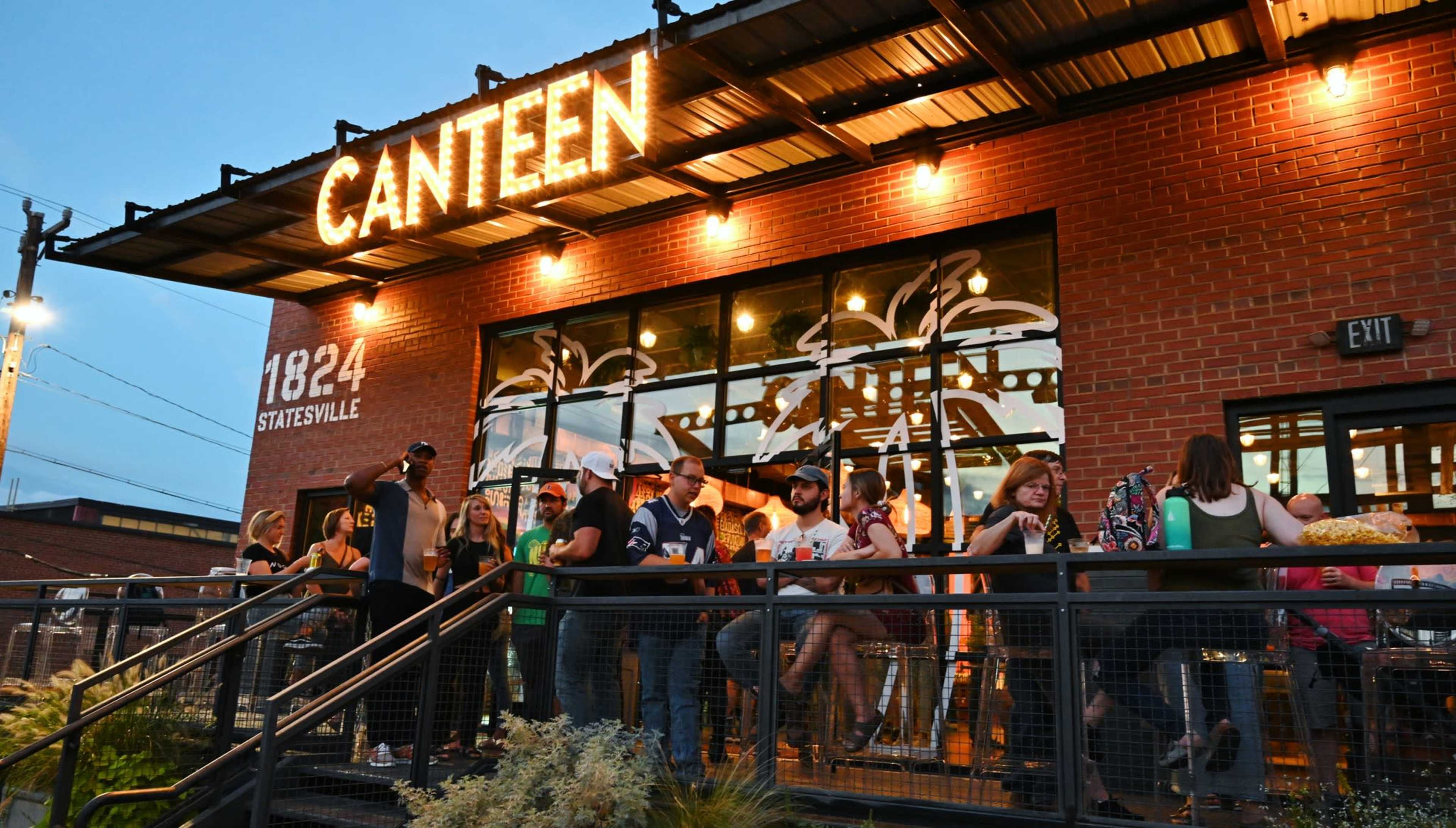 A crowd gathers outside the Canteen restaurant, with people sitting at tables and mingling on the patio as evening approaches.