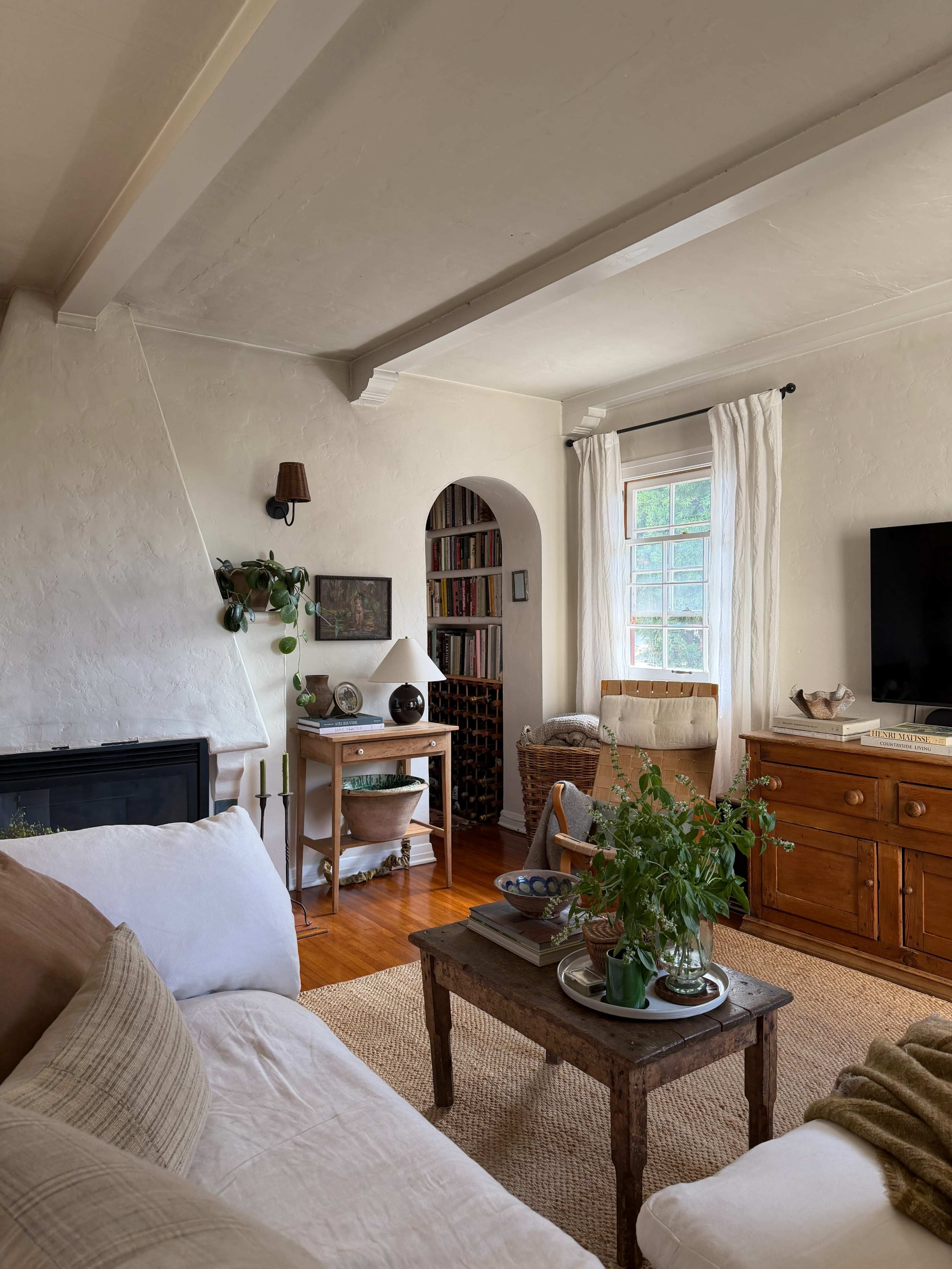 The interior of a cozy living room features a white sofa, a wooden coffee table, and a small dining area, with bookshelves and natural light streaming through a window.