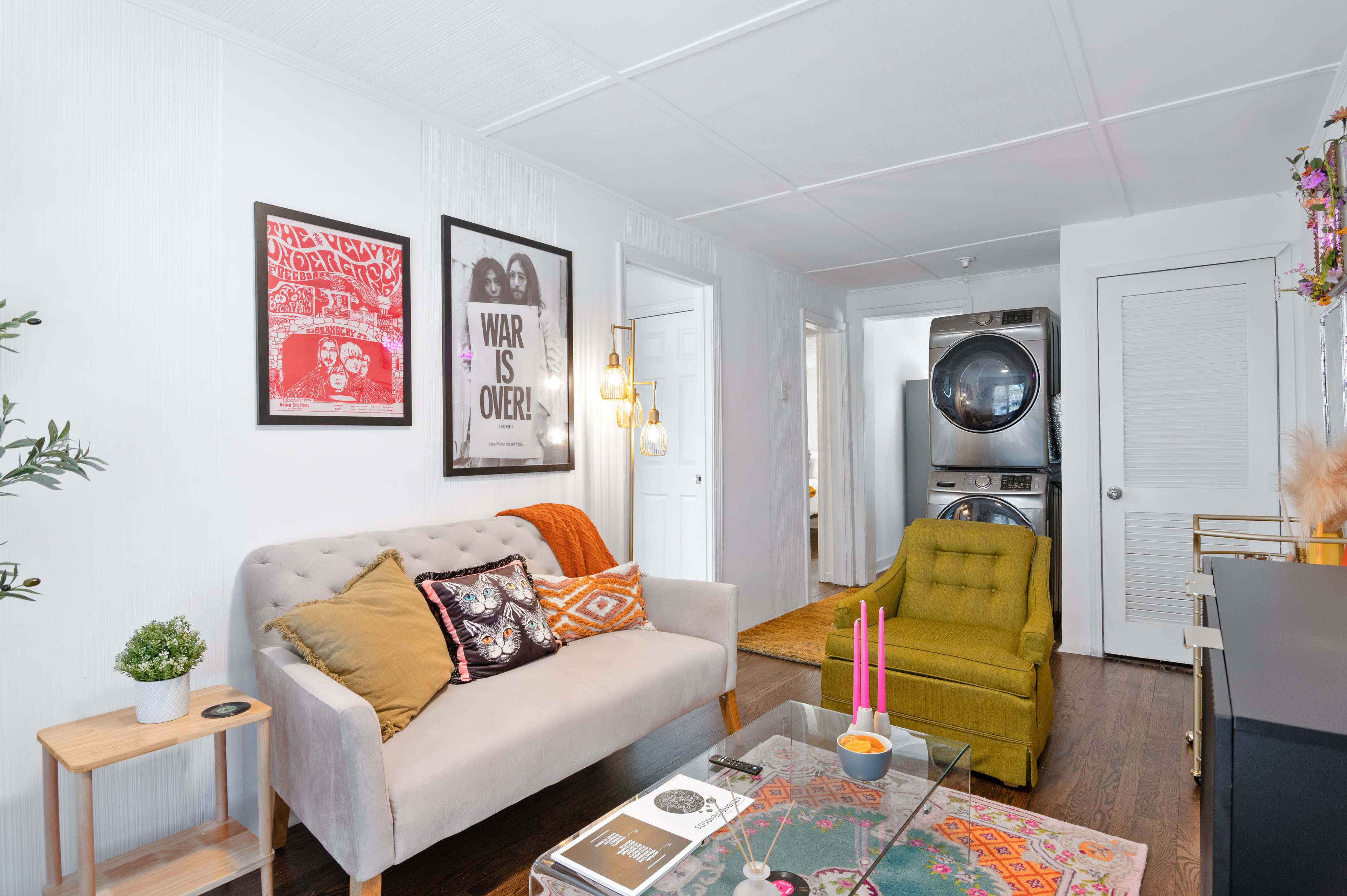 A living room with a gray sofa, a green chair, a coffee table with decorative items, and framed artwork on the walls, along with a laundry area in the background.