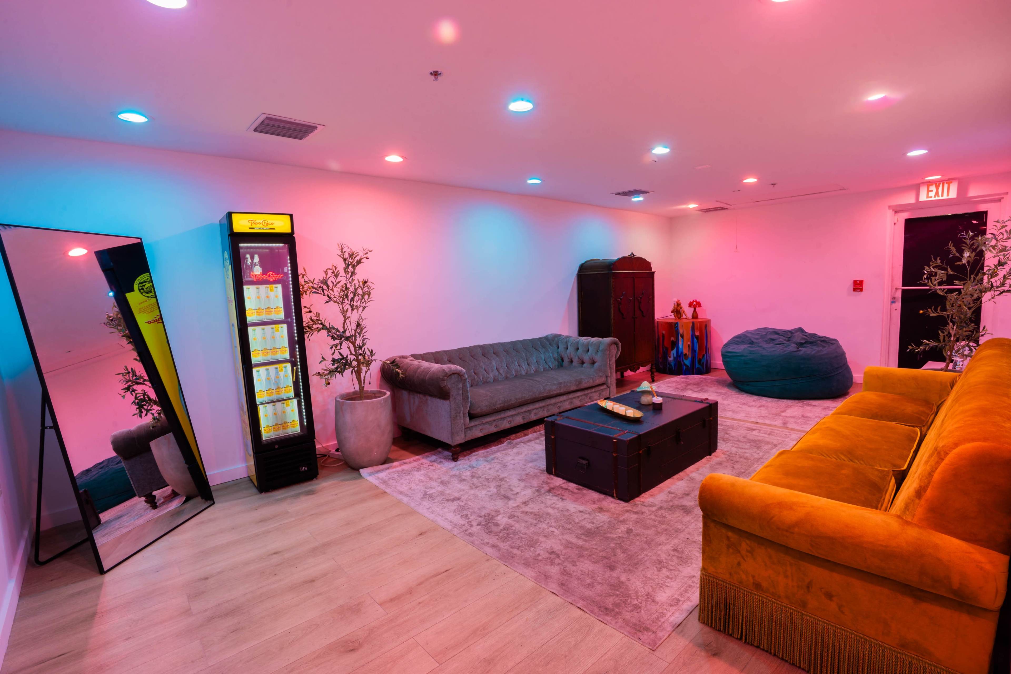 The scene features a brightly lit lounge area with two sofas, a coffee table, a mirror, and a vending machine, all arranged on a plush rug.