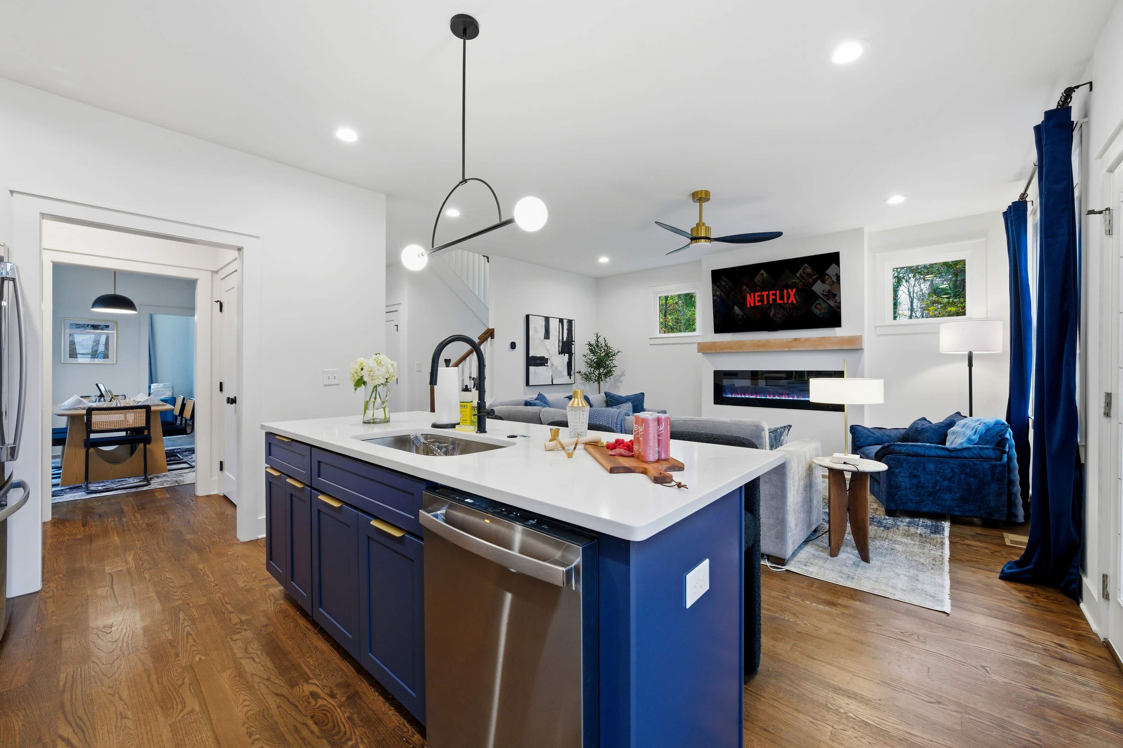 A modern kitchen opens to a living room, featuring a central island with a sink and a view of a cozy seating area with a fireplace and a television.