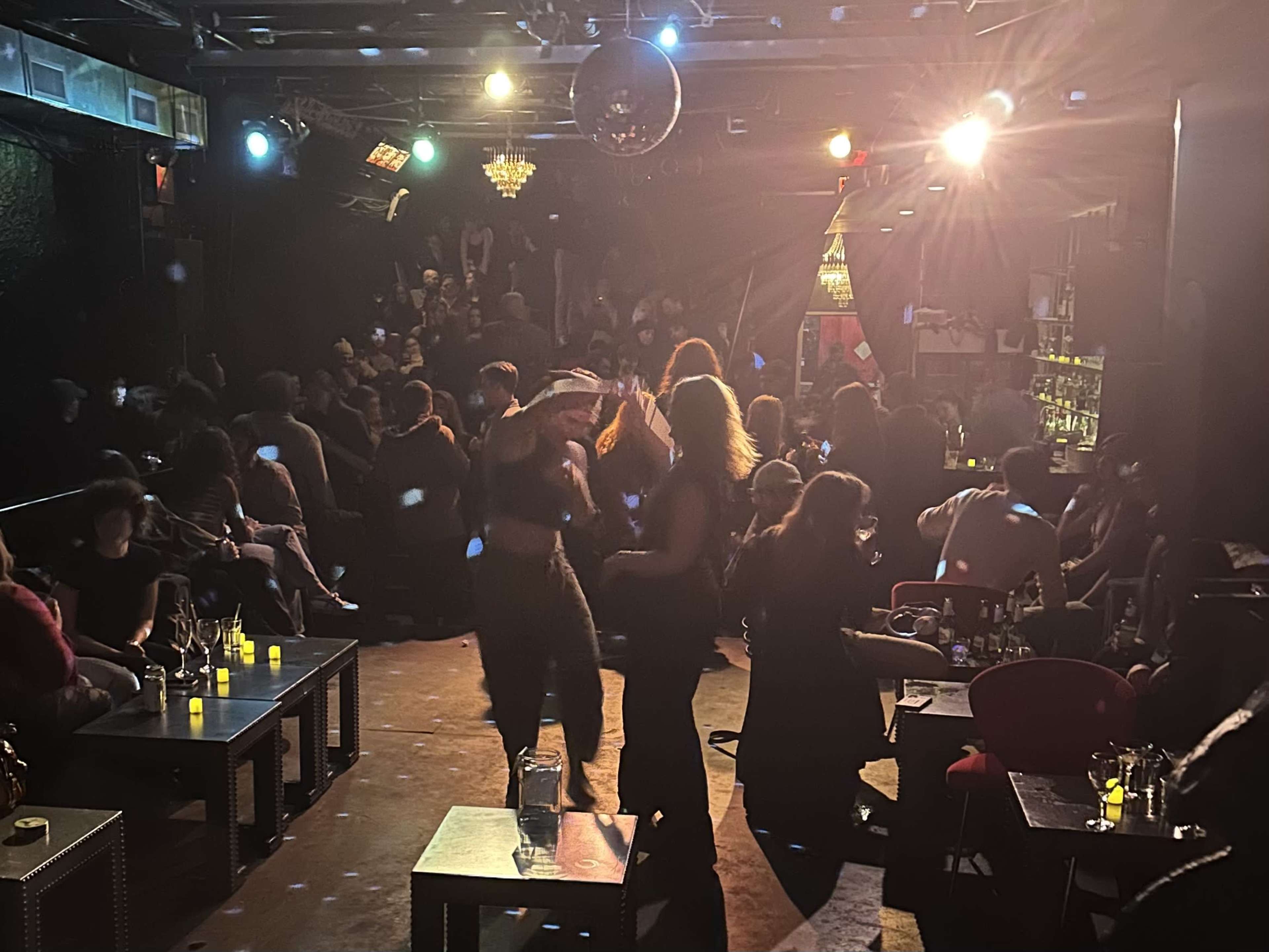 A dimly lit nightclub scene shows a crowd of people socializing and dancing near a bar, with disco lights illuminating the space.