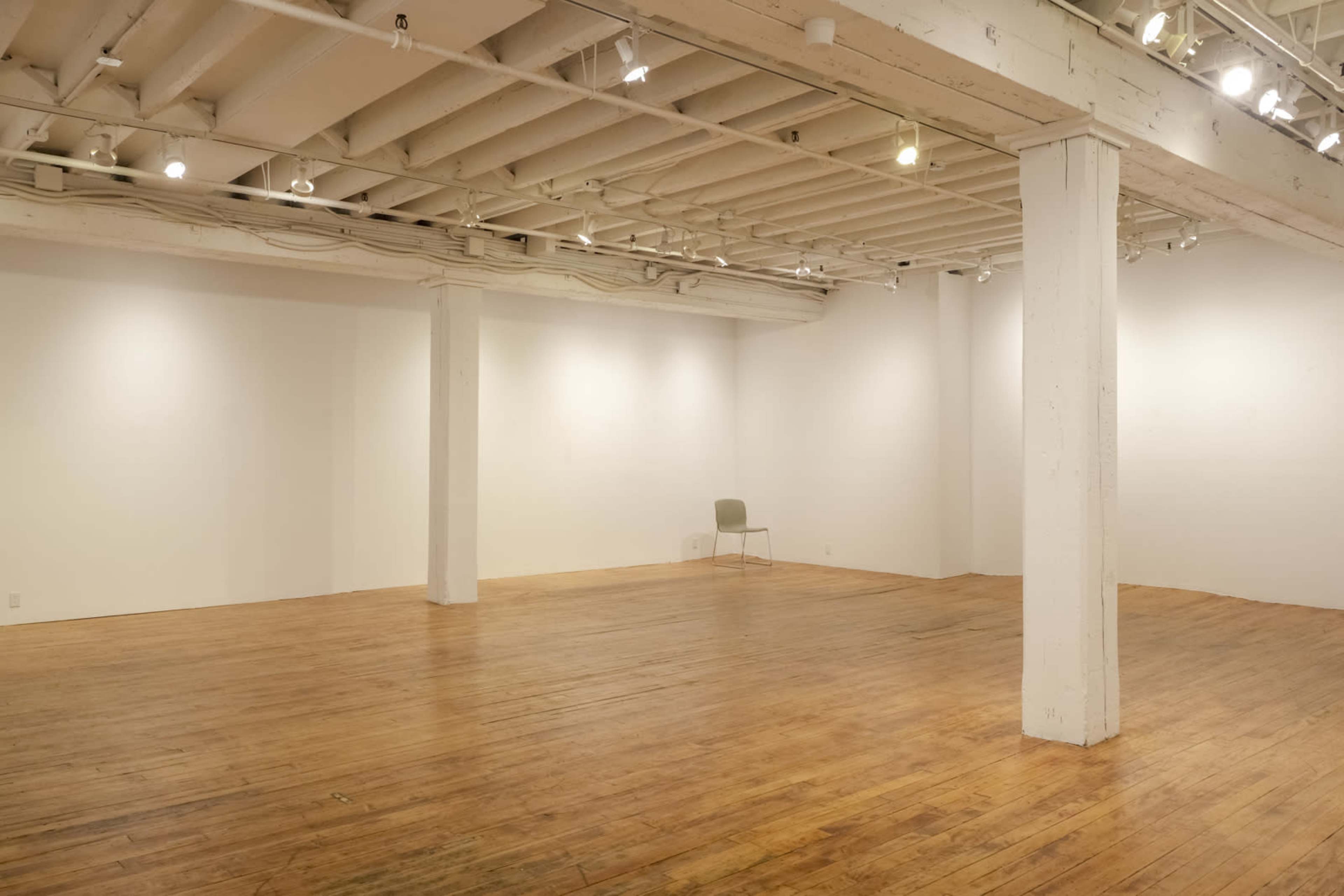 The image shows a spacious, empty gallery with wooden flooring, white walls, and a single chair positioned in the center.