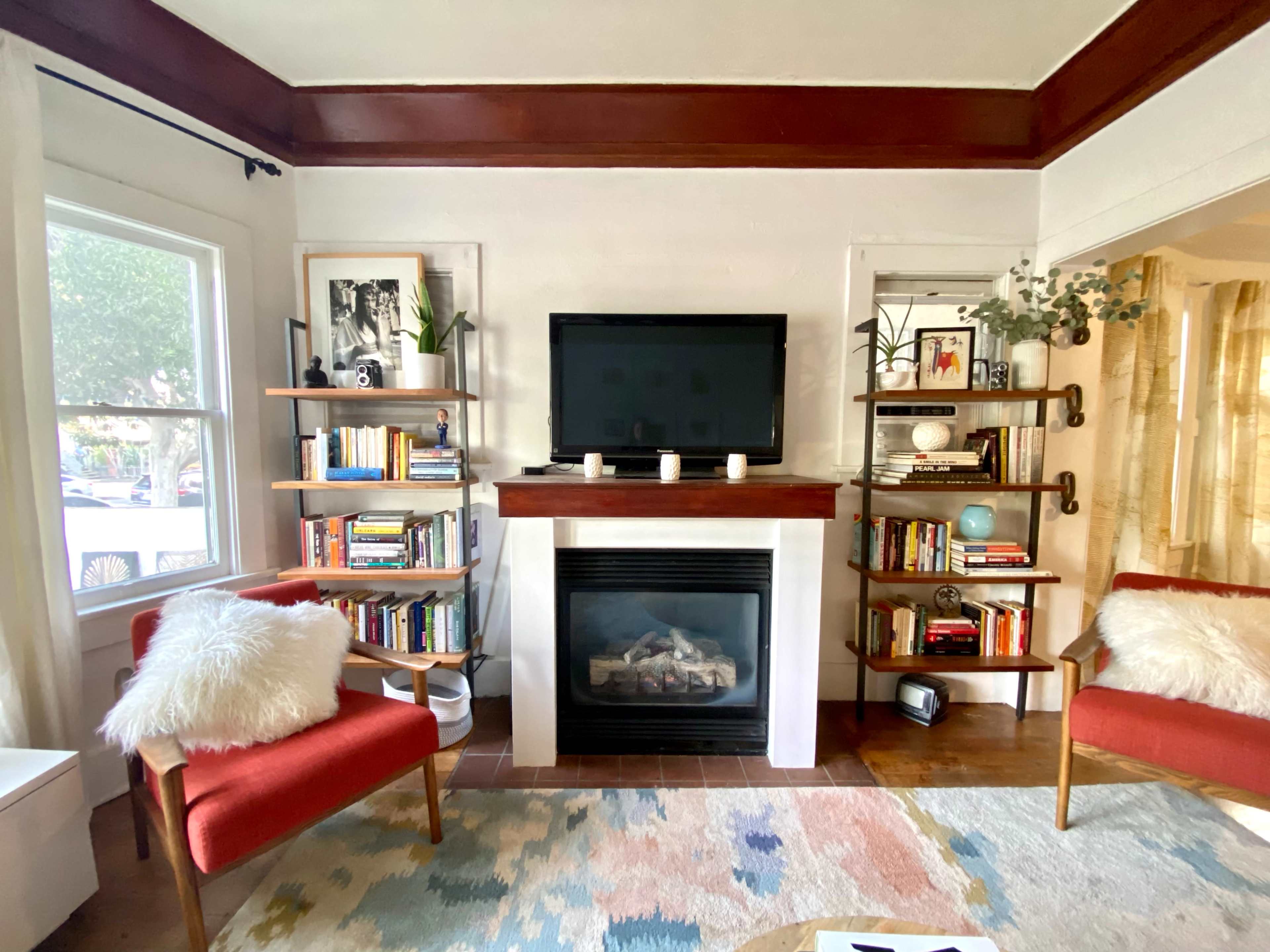The image shows a living room with a fireplace, bookshelves filled with books, two red armchairs, and a television mounted on the wall.