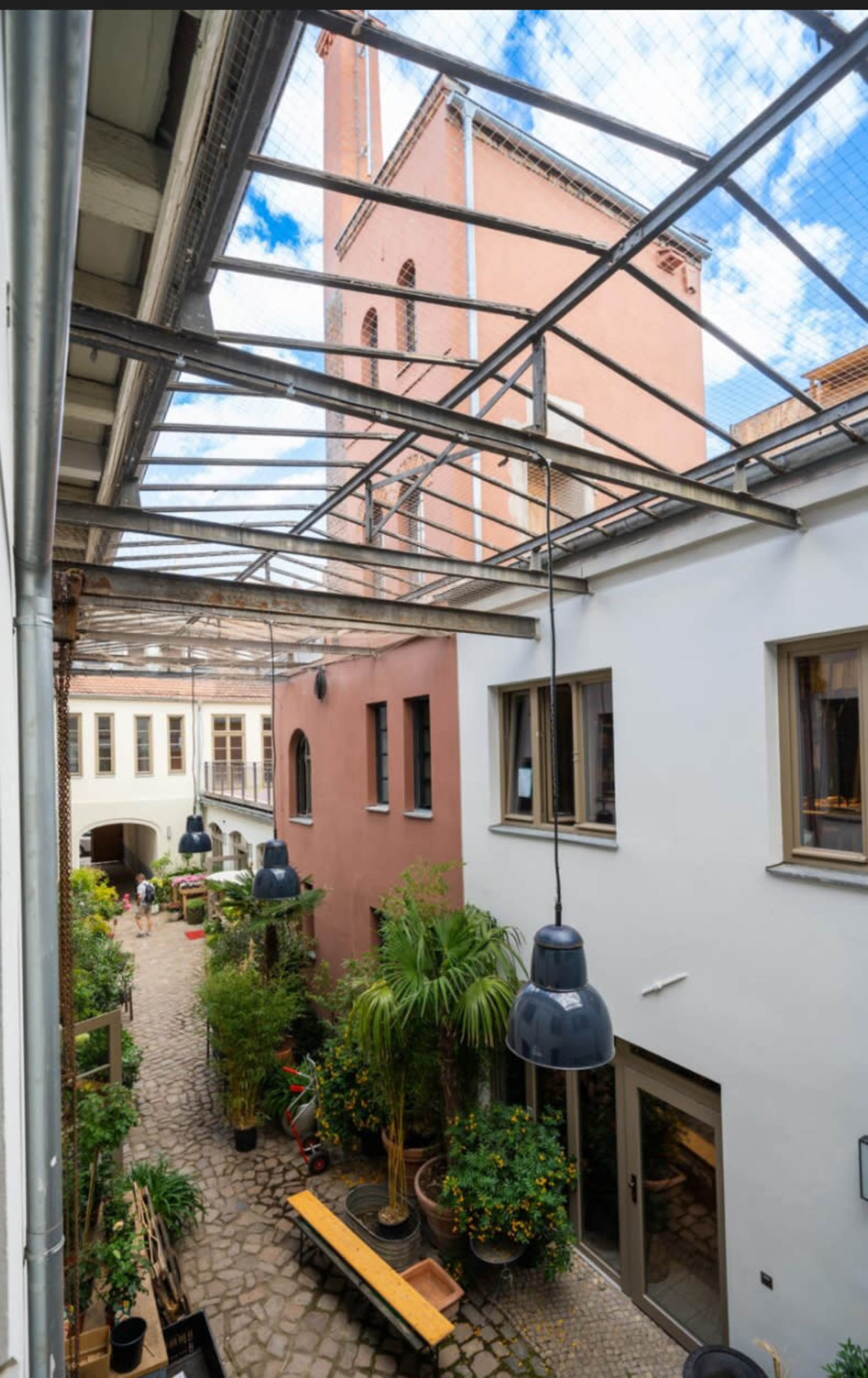 The image shows a courtyard surrounded by buildings with a clear glass roof, featuring potted plants, a wooden bench, and hanging lights above a cobblestone path.