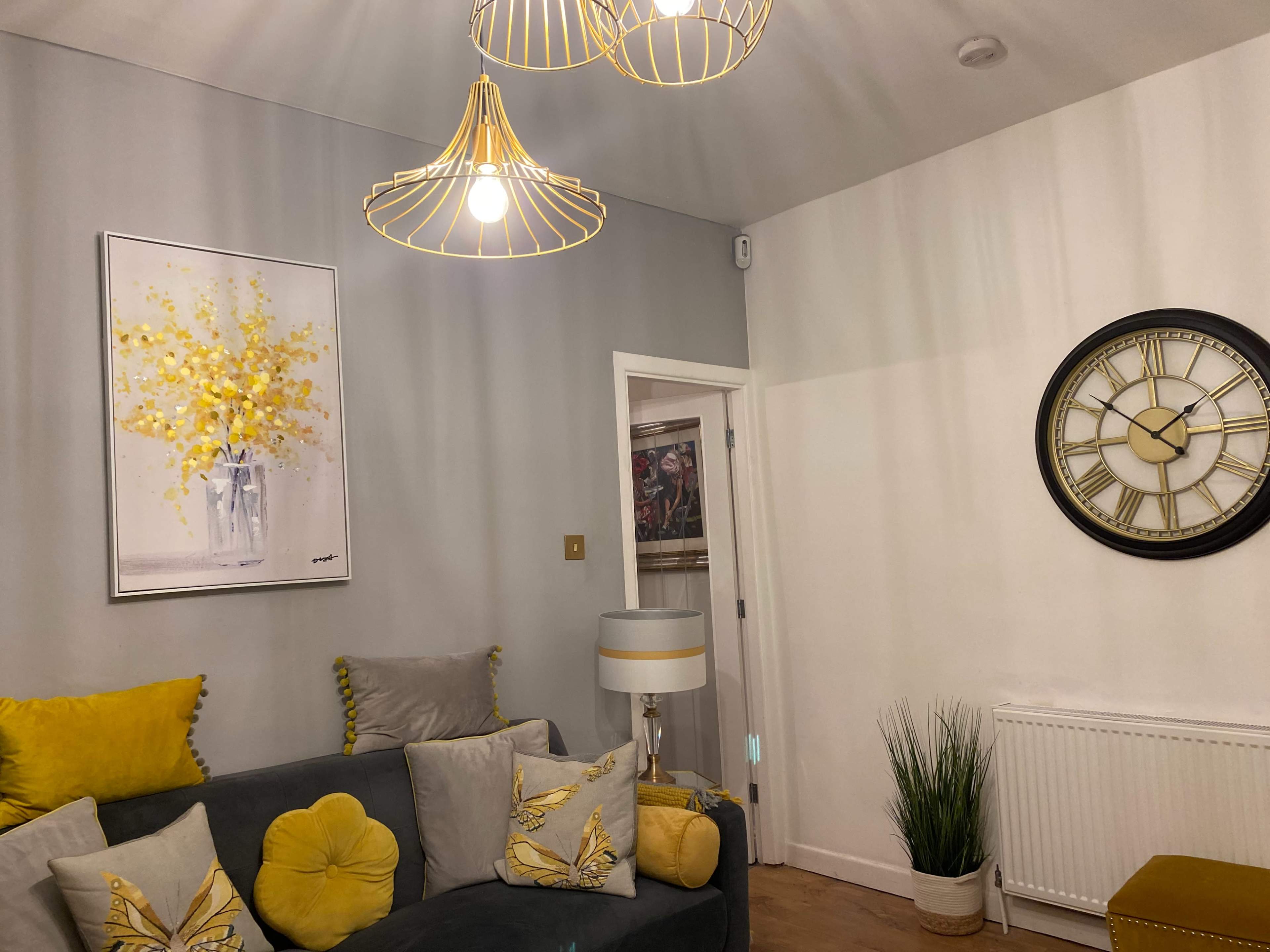 A living room with a grey wall, yellow accent decorations, a large wall clock, and a floral painting.