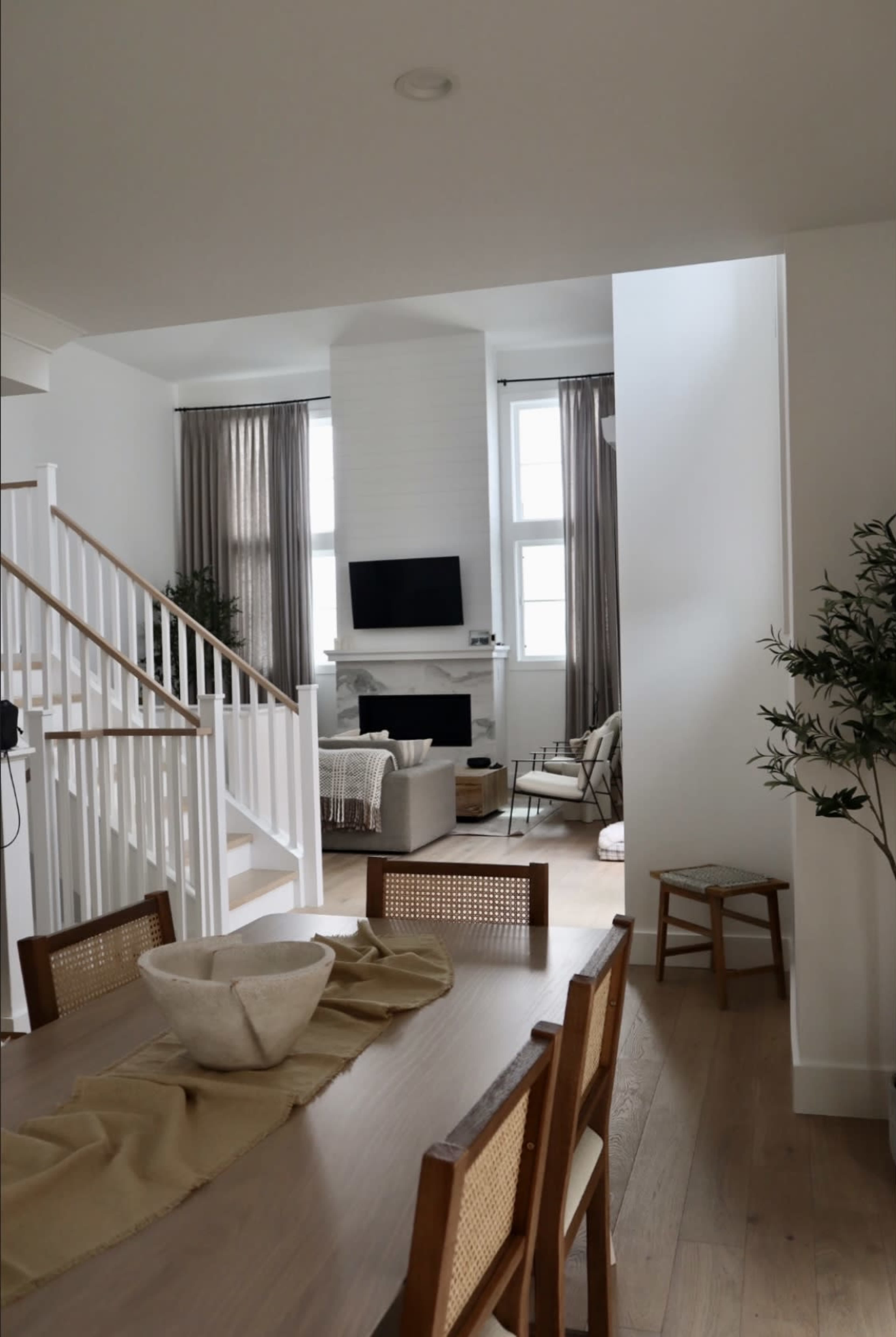 A modern, open-concept interior features a dining area with wooden chairs, a table set with a bowl, and a living space beyond that includes a fireplace and large windows.