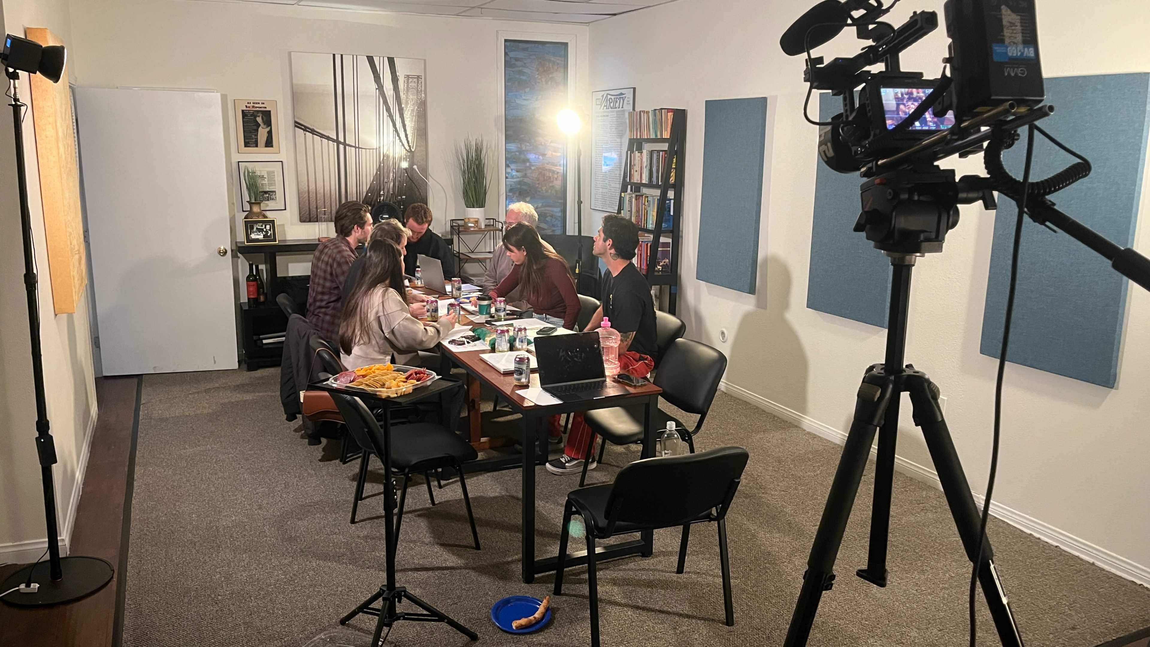 A group of six people is seated around a table in an office-like setting, engaged in a discussion while various items are spread out on the table.