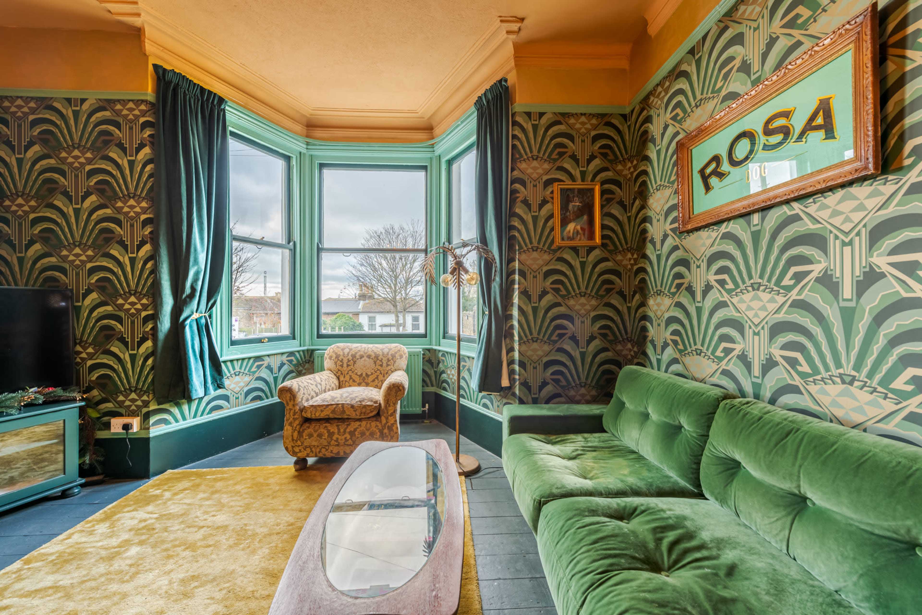 A room with green patterned wallpaper, a green velvet sofa, a vintage armchair, and a large window with decorative curtains.