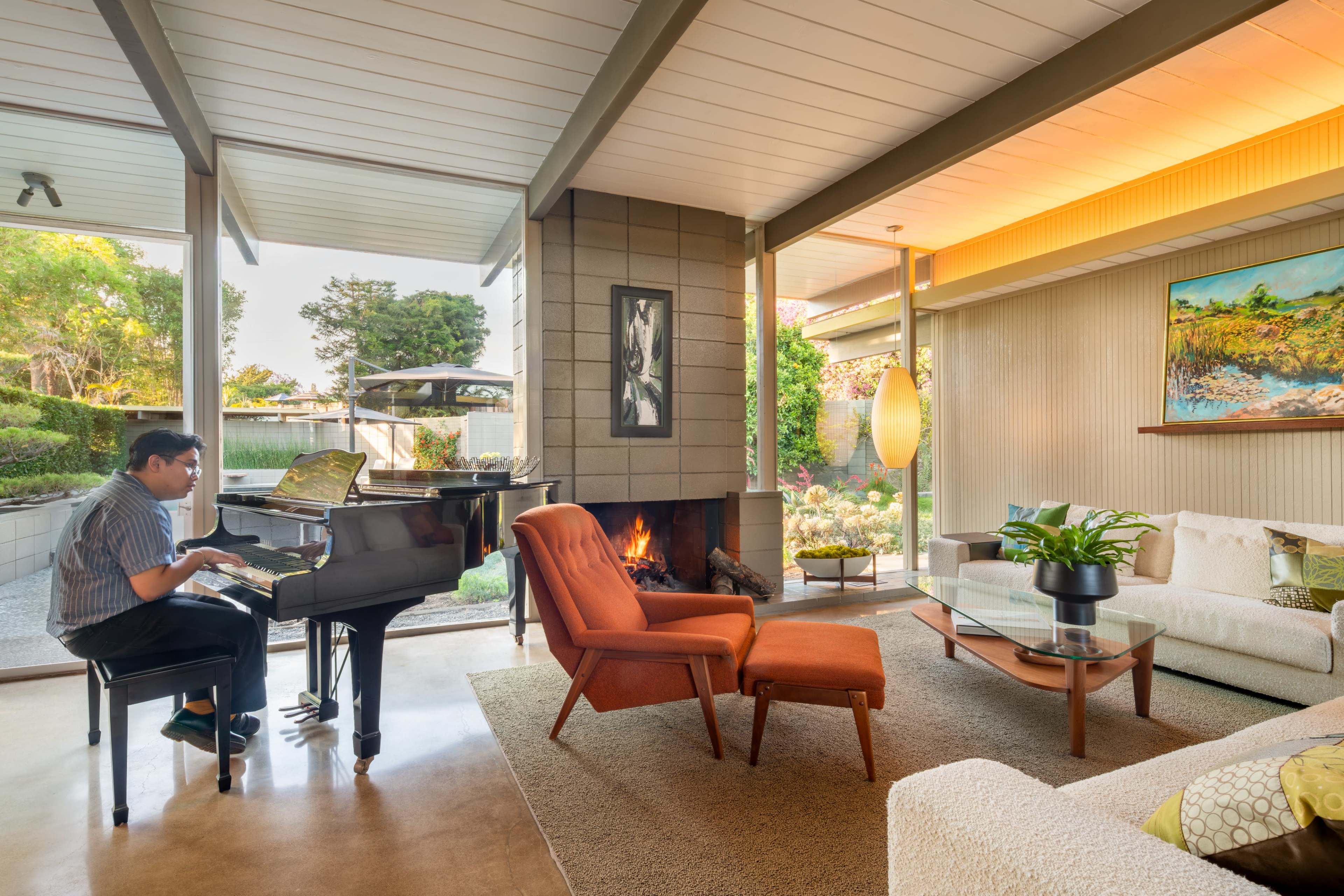 A person plays a piano beside a modern living room featuring a cozy fireplace and large windows that open to a garden.