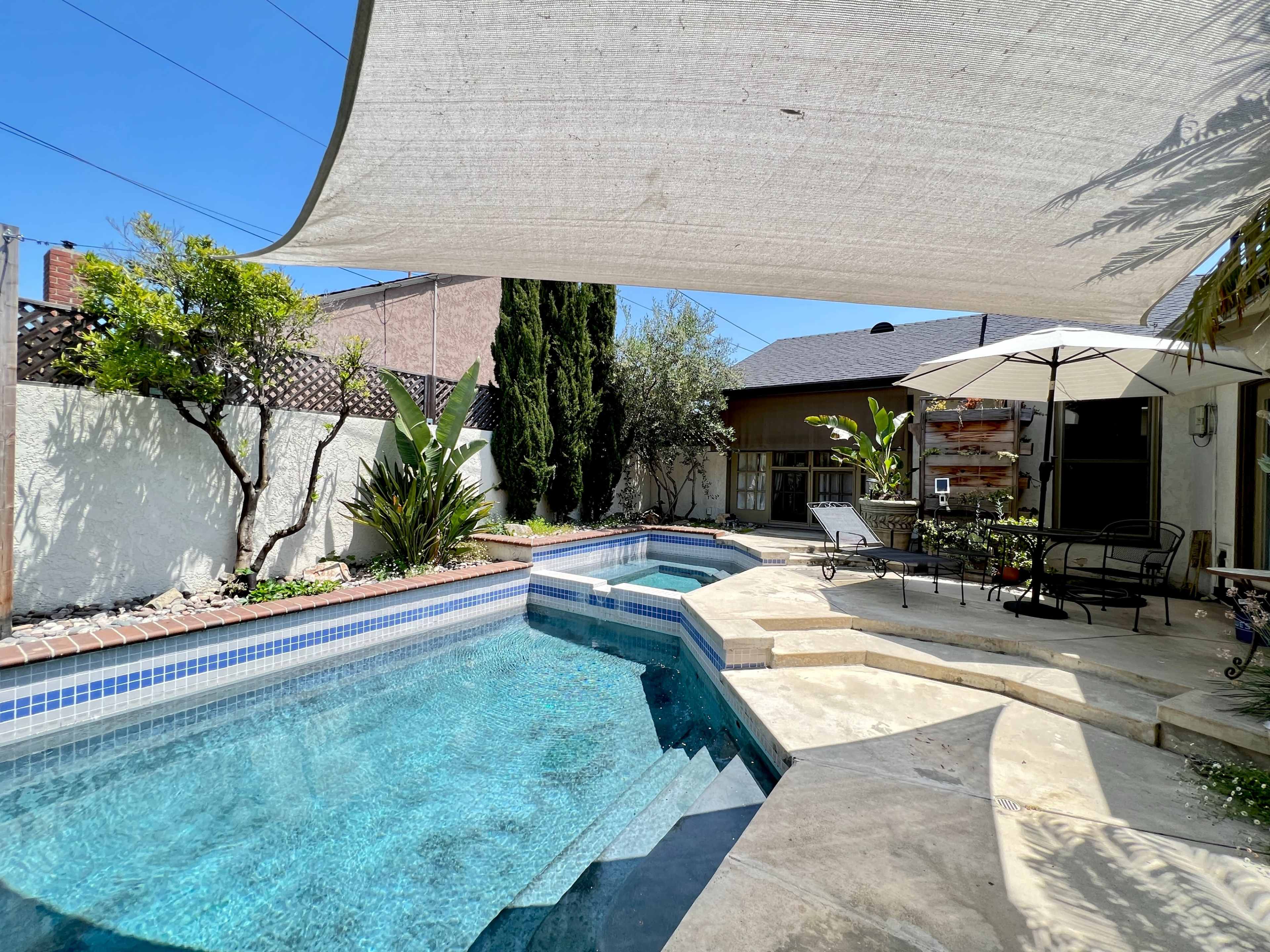 The image shows a backyard with a swimming pool, spa, shaded seating area, and surrounding greenery.