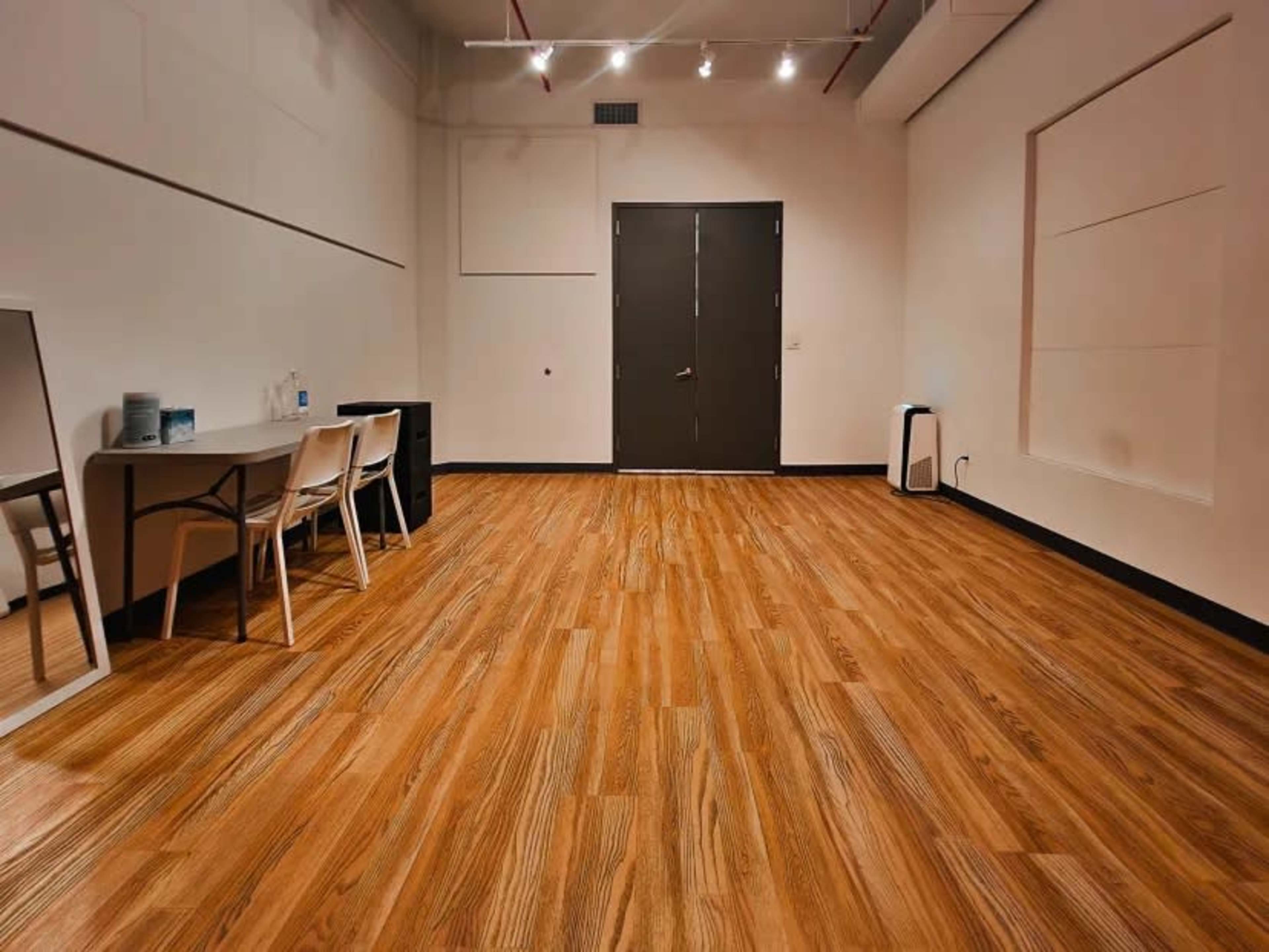 The image shows a simple, empty room with wooden flooring, a table with chairs, a mirror, and a closed door.