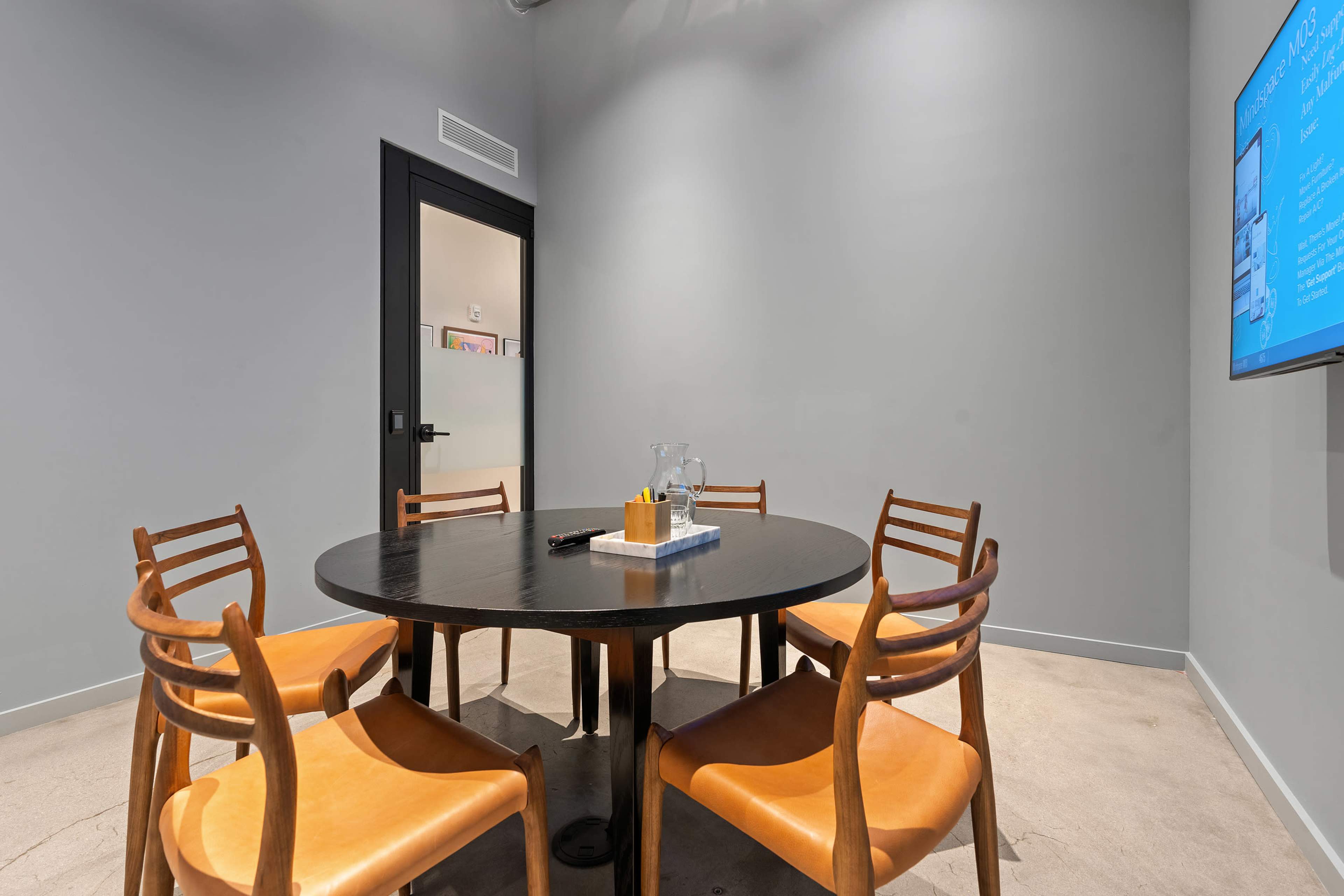 A small conference room features a round black table surrounded by four wooden chairs, with a glass pitcher and a notepad on the table.