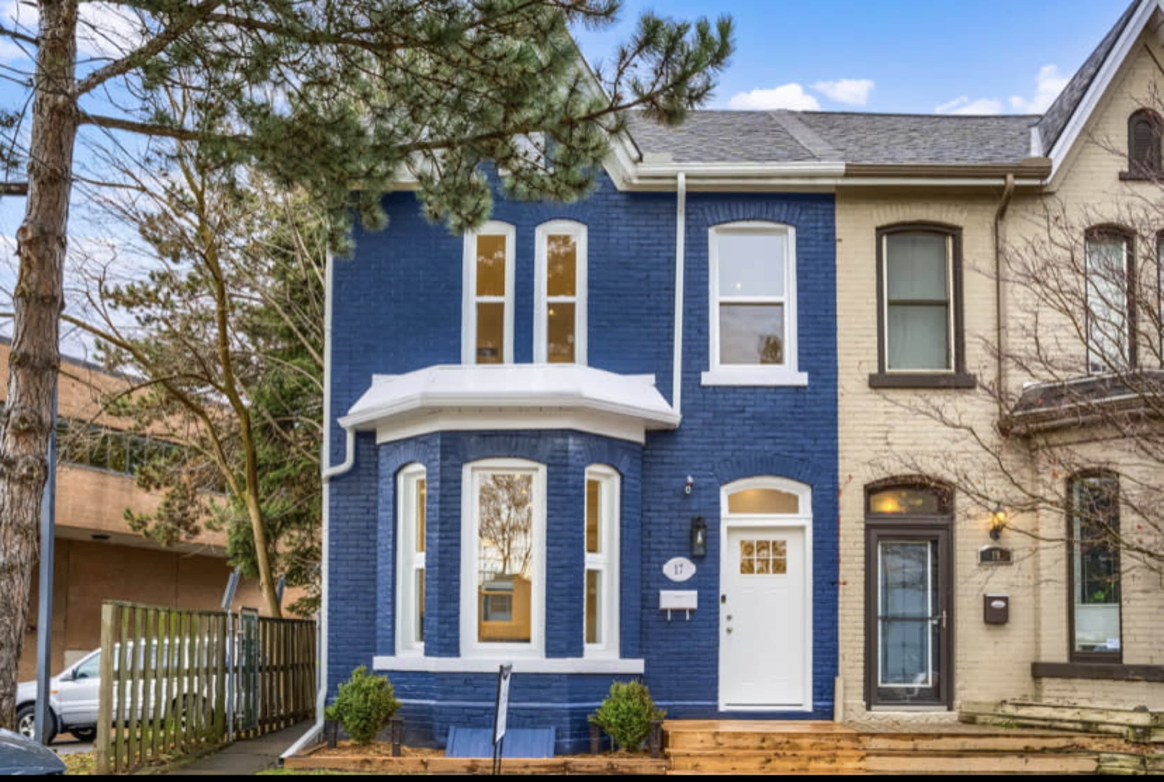 A two-story brick townhouse with a blue facade and white trim, situated next to a light-colored neighboring building and featuring a wooden front porch.
