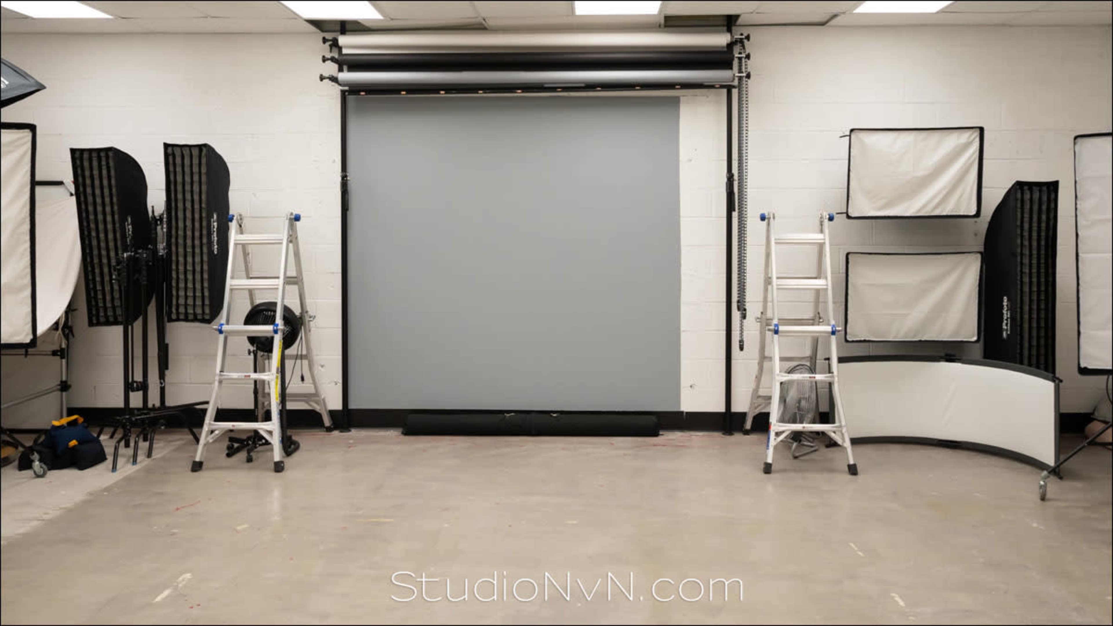 A photography studio is set up with a gray backdrop, two step ladders, and various light modifiers arranged along the walls.